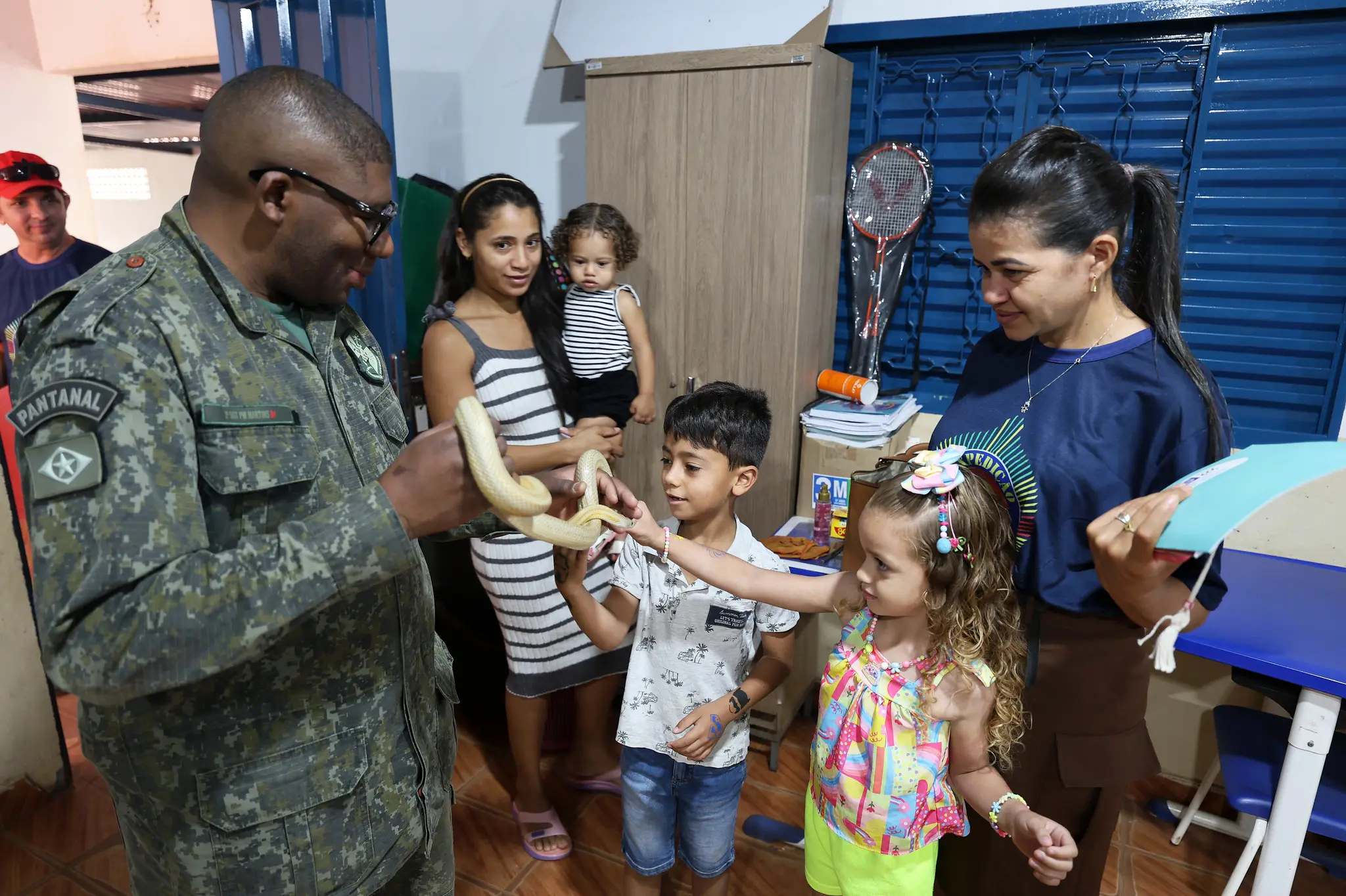 Policial militar com roupa verde camuflada, segurando cobra de clara, com tons amarelados e apresentando animal a crianças, durante atividade de educação ambiental.