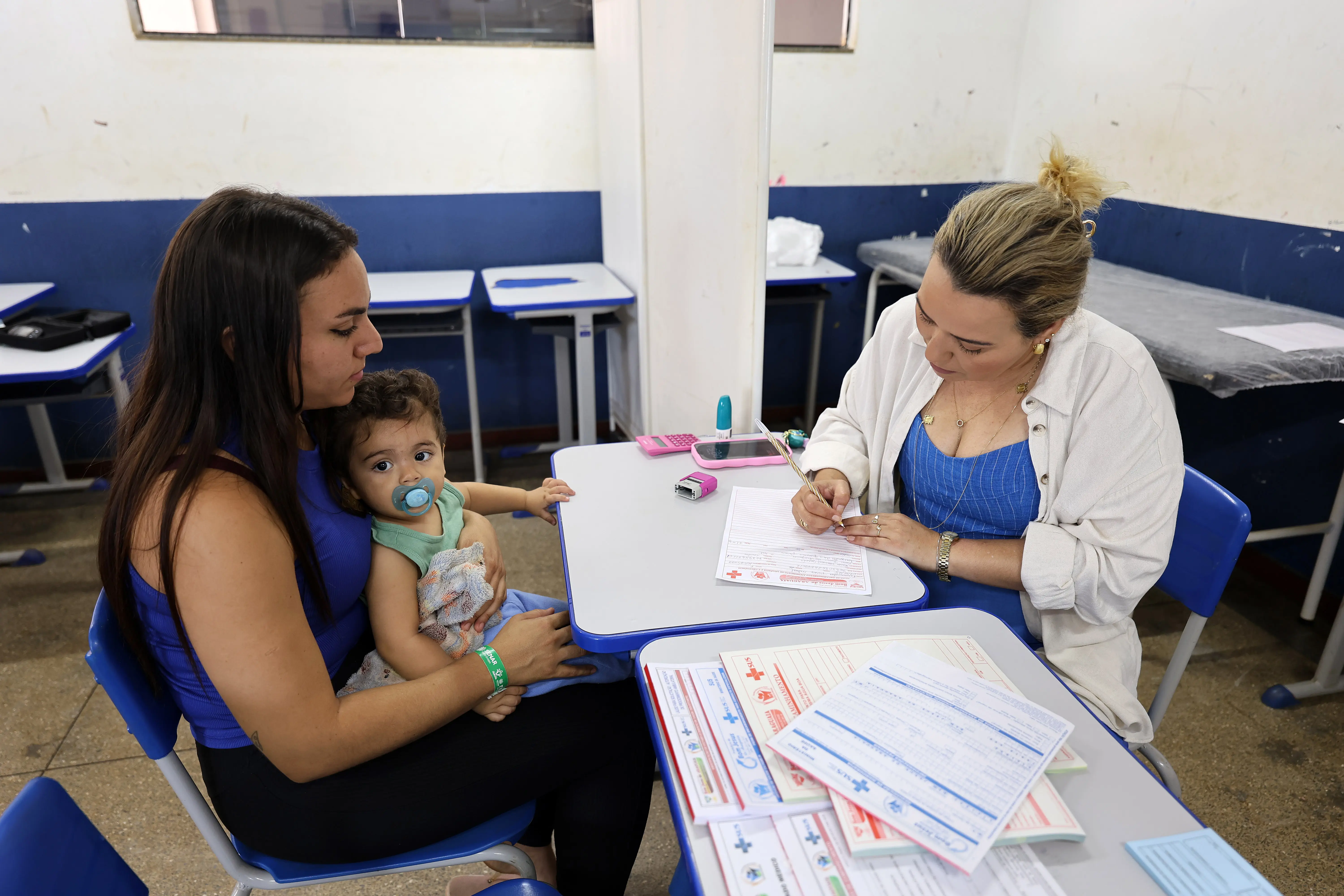 Mulher segura um bebê no colo enquanto outra profissional preenche formulários sobre uma mesa. A cena ocorre em sala simples, mostrando atendimento humanizado e acolhimento durante ação social.