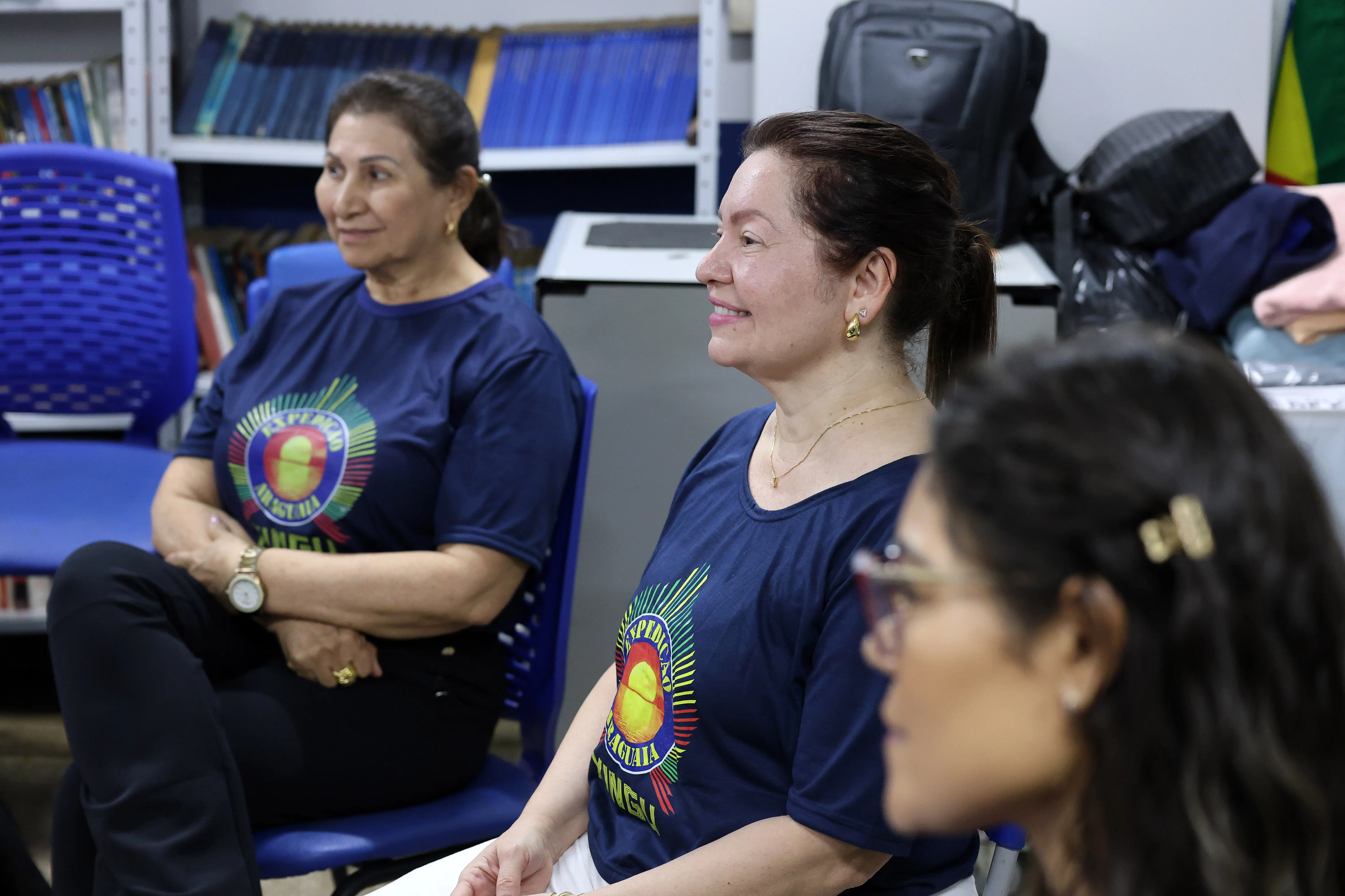 Três pessoas estão sentadas em cadeiras azuis, usando camisetas com logotipo colorido. Ao fundo, estantes com livros e uma mochila sobre a mesa indicam um ambiente educacional, como sala de aula ou biblioteca.