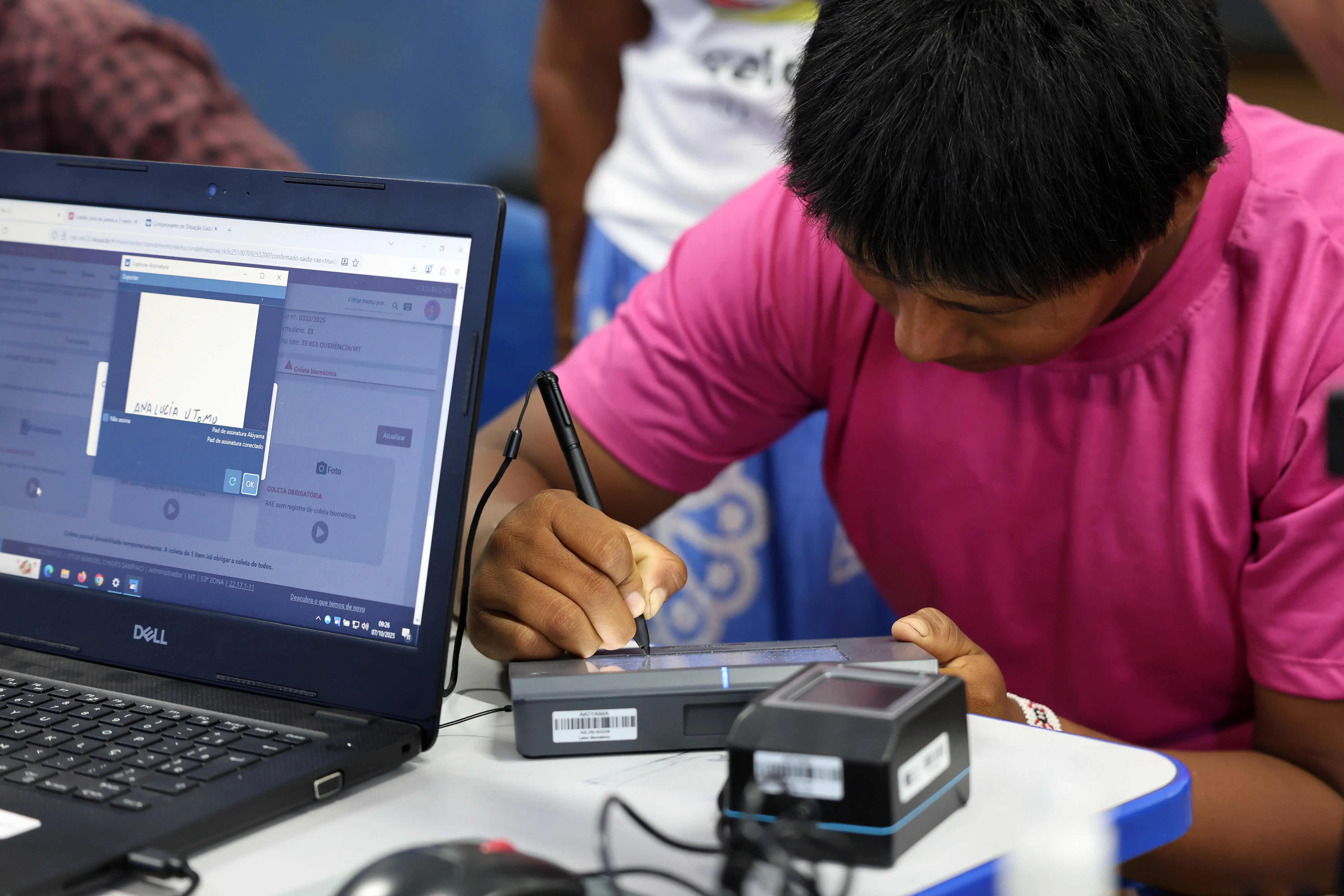 Jovem indígena Xavante, de blusa rosa, assina documento digital em mesa com notebook e equipamento biométrico durante atendimento da Expedição Araguaia-Xingu. O ambiente é simples, com parede azul e clima de concentração.