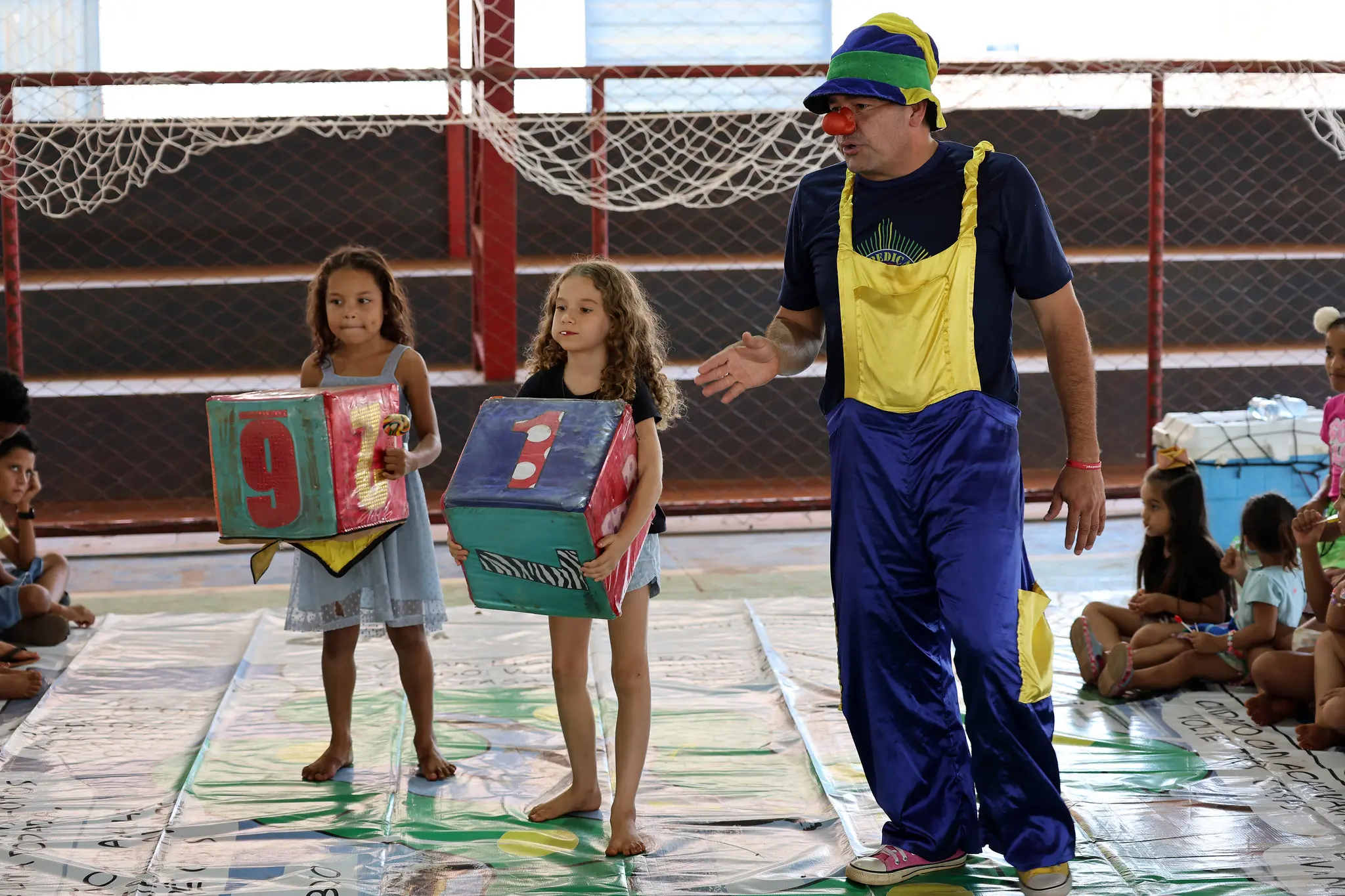 Marcelo Luciano, policial vestido de palhaço, com roupa azul, amarela e verde e chapéu das mesmas cores, em atividade recreativa com crianças, que seguram dados sobre a lona do jogo Rebojando, durante a Expedição Araguaia-Xingu.