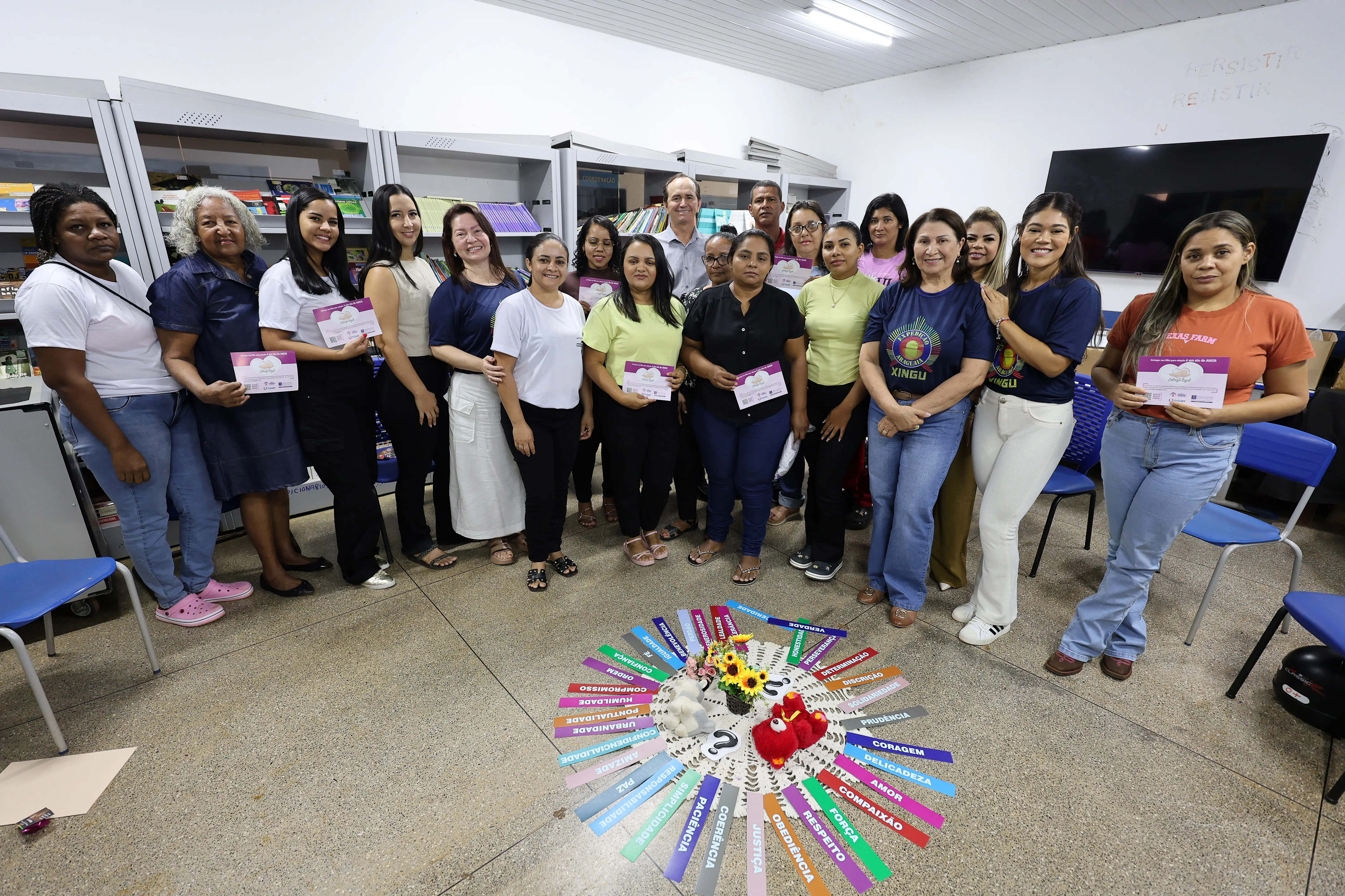 Grupo de pessoas em pé, posando com folhetos informativos. Estão organizados ao redor do arranjo colorido. A sala continua sendo a biblioteca, com atmosfera de integração e conscientização.