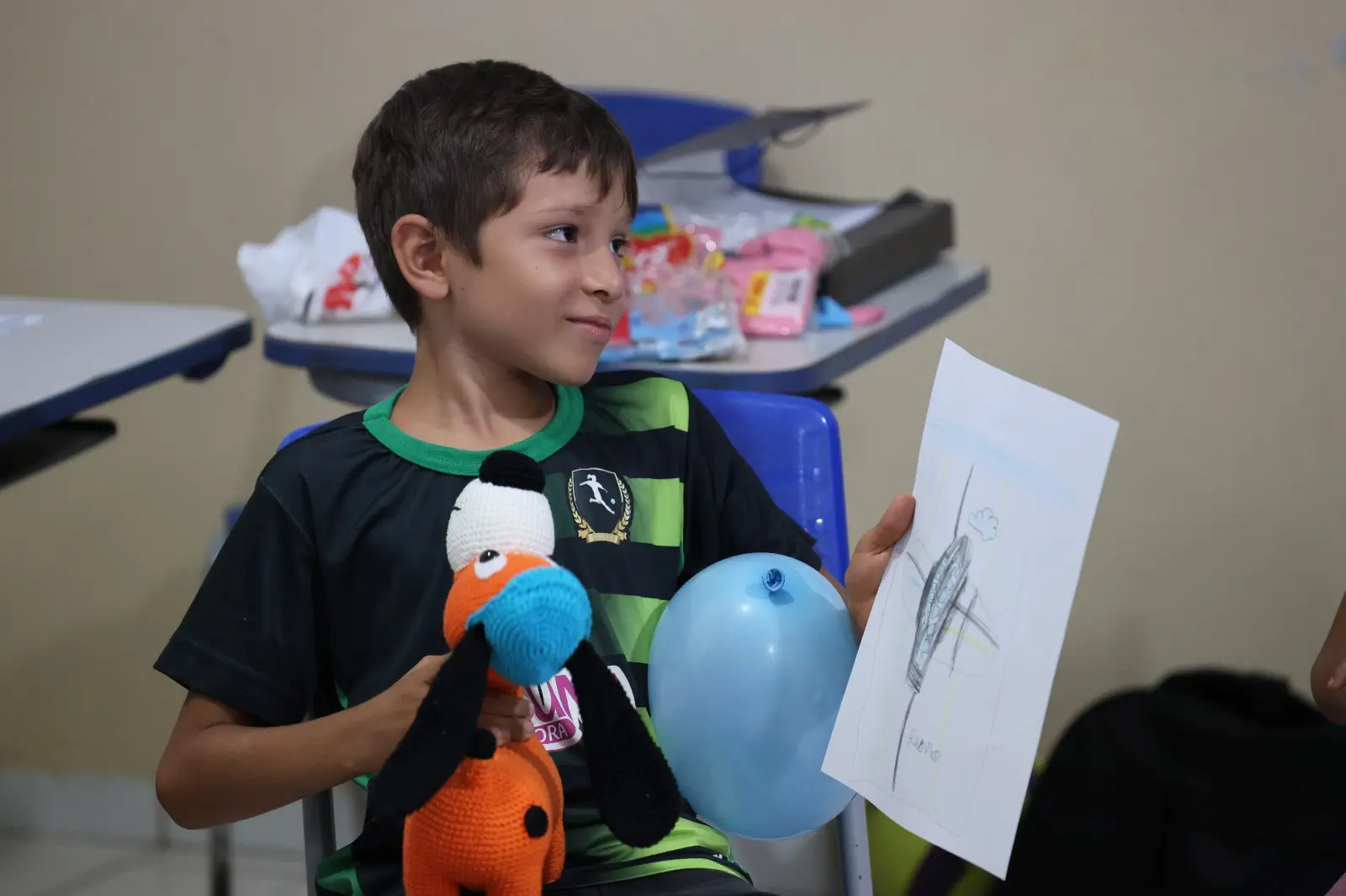 Menino segura balão azul, desenho e brinquedo de crochê enquanto sorri discretamente. O ambiente mostra mesas com materiais coloridos e clima de leveza