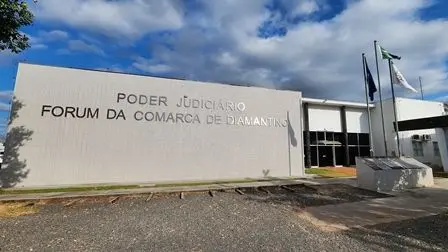 Fachada do Fórum da Comarca de Diamantino, em Mato Grosso, com o letreiro “Poder Judiciário – Fórum da Comarca de Diamantino” em destaque na parede branca. À direita, há três mastros com bandeiras hasteadas e a entrada principal do prédio, composta por portas de vidro. 