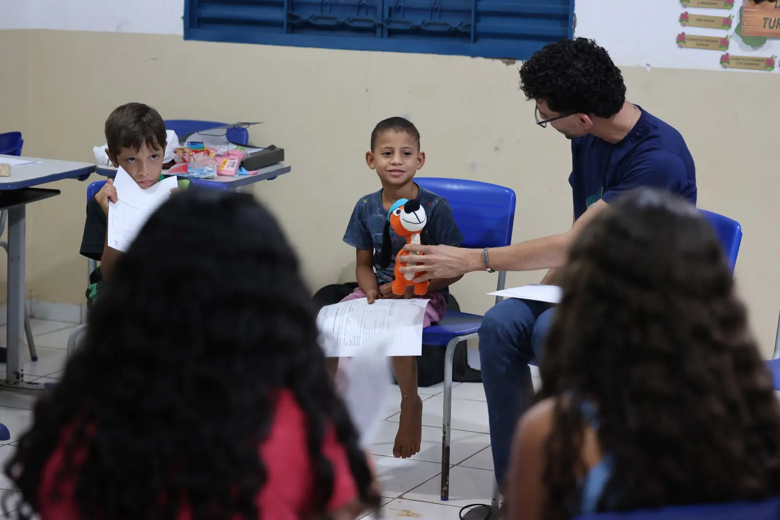 Educador segura um brinquedo de crochê e conversa com menino durante a roda de diálogo. As crianças participam com interesse, segurando folhas e desenhos.