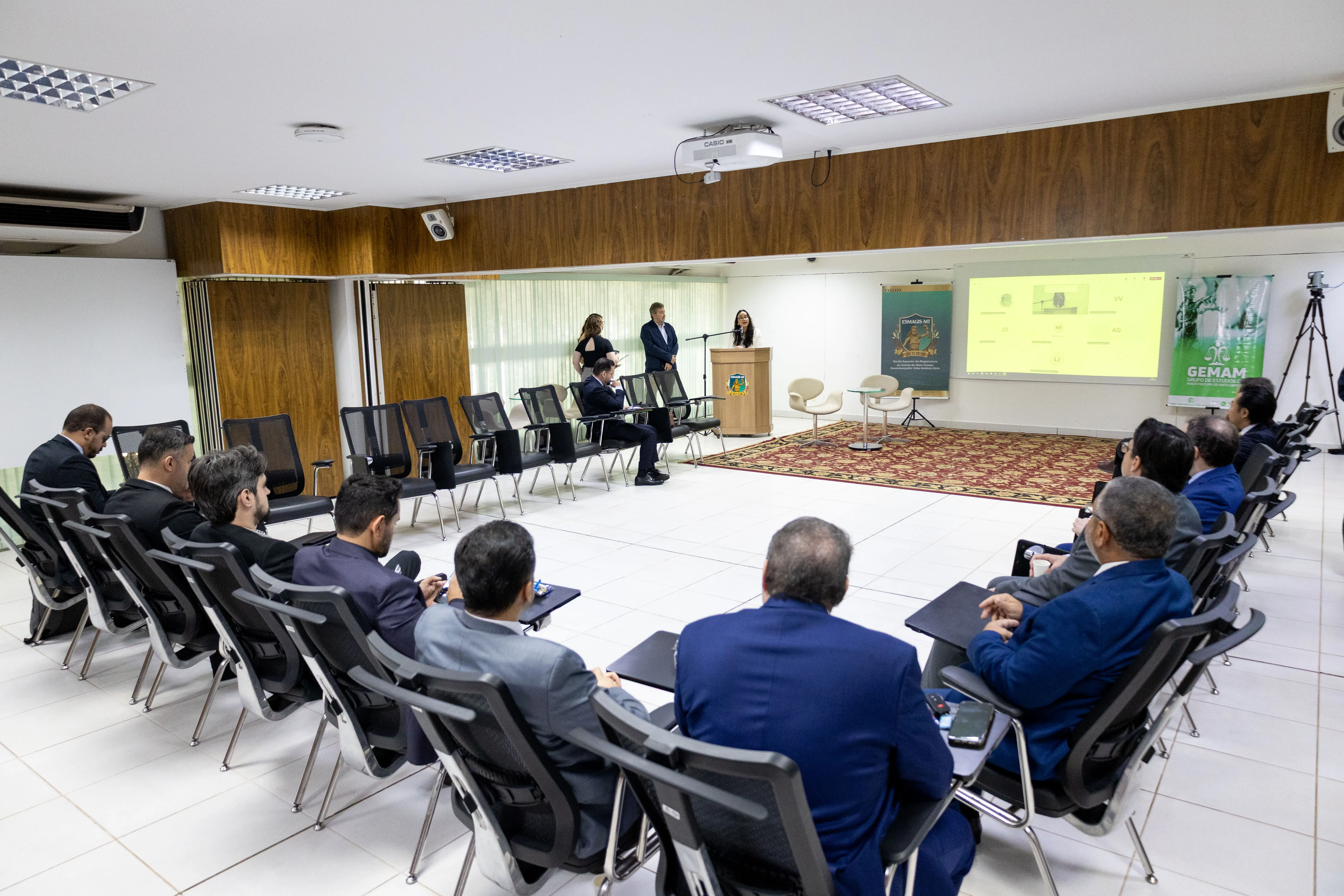 Sala de conferência com público em semicírculo. Duas pessoas apresentam conteúdo projetado em tela. Ambiente tem paredes brancas, detalhes em madeira e banners com destaque para “GEMAM”.
