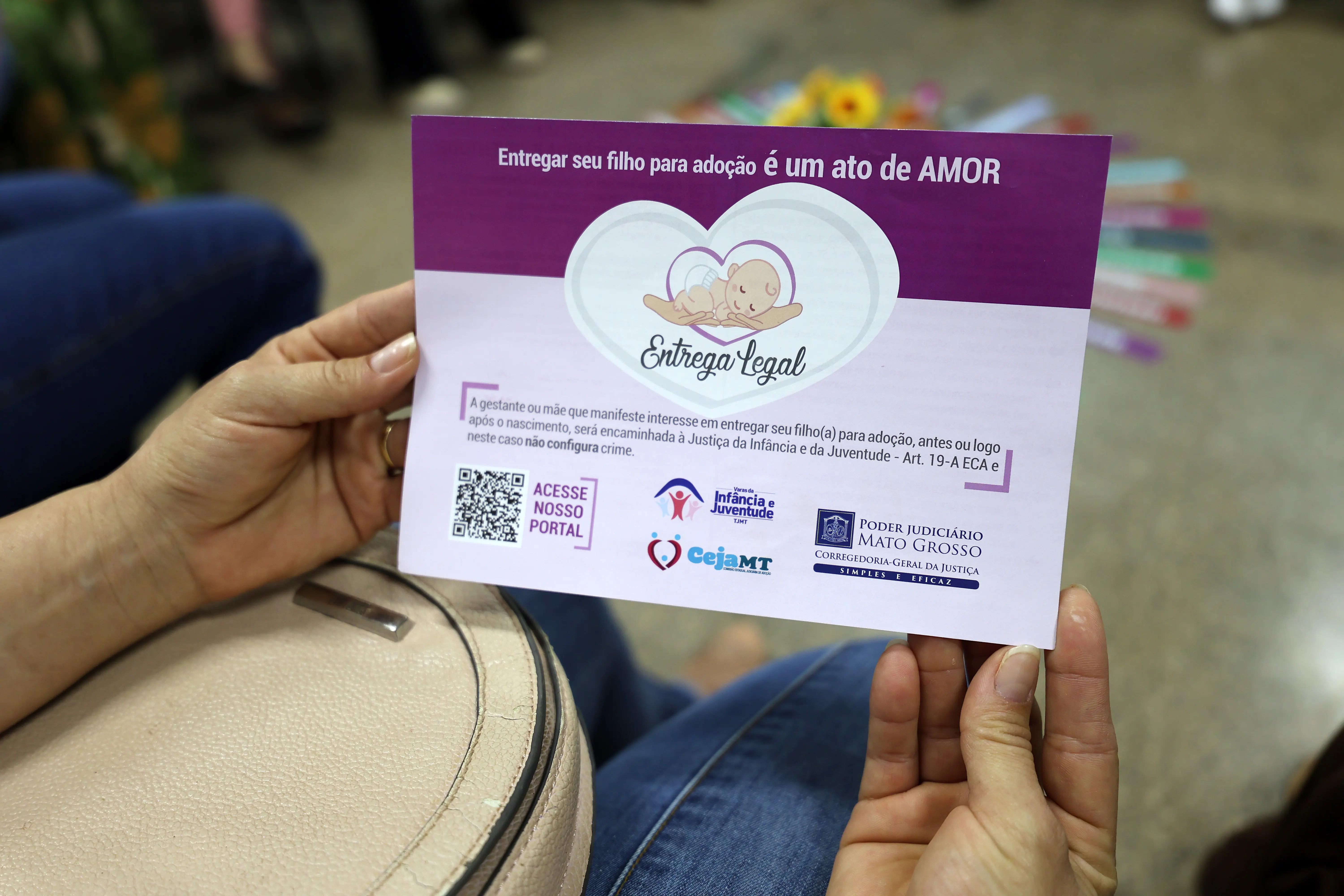 Pessoa segura panfleto com mensagem: “Entregar seu filho para adoção é um ato de AMOR”. O material promove a entrega legal, com contatos e QR codes. O foco é educativo e voltado à conscientização social.
