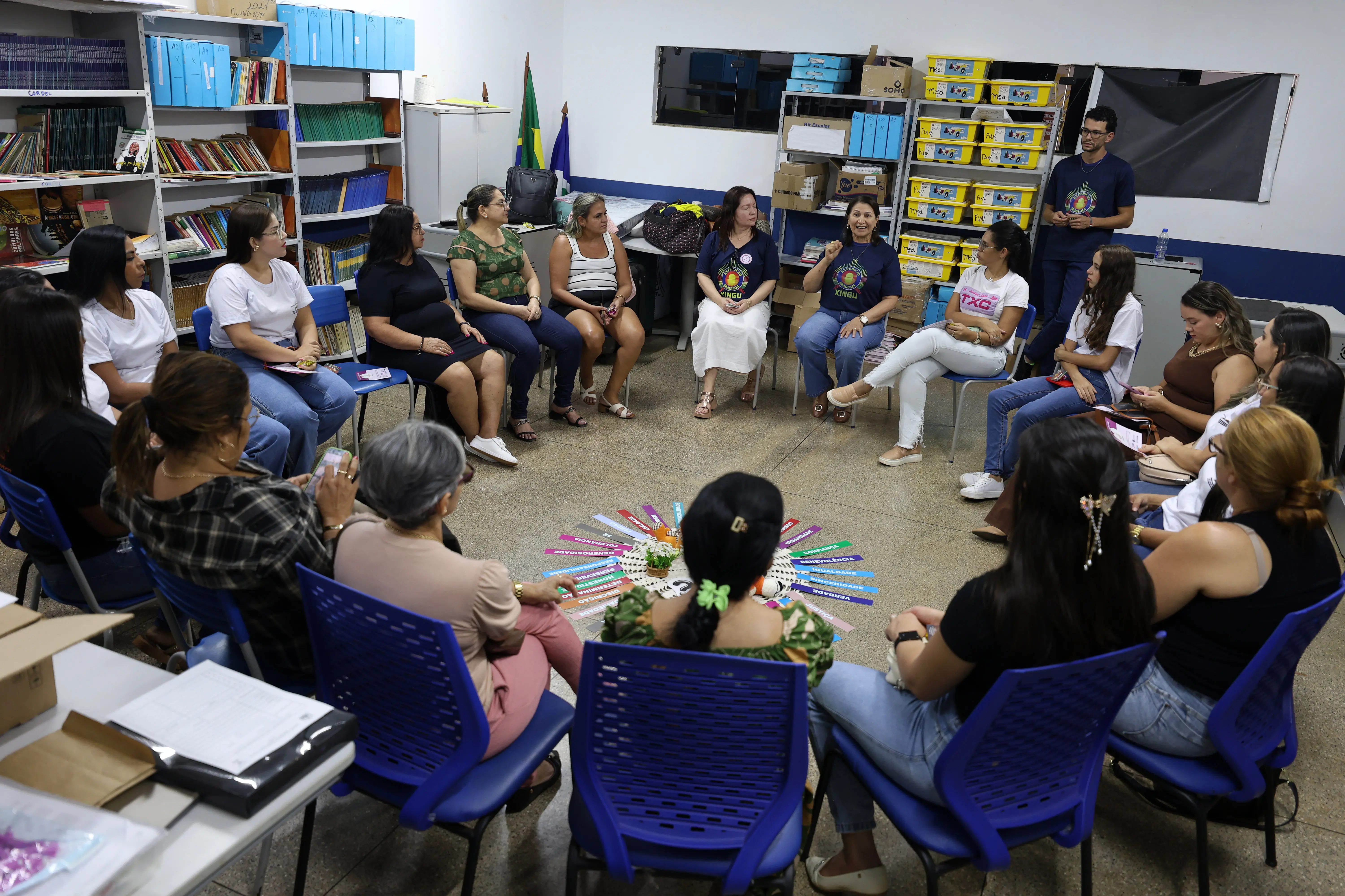 Sala ampla com grupo em círculo. Pessoas sentadas em cadeiras azuis, cercadas por estantes com livros e caixas. No centro, objetos coloridos dispostos radialmente sugerem dinâmica coletiva ou atividade de integração.