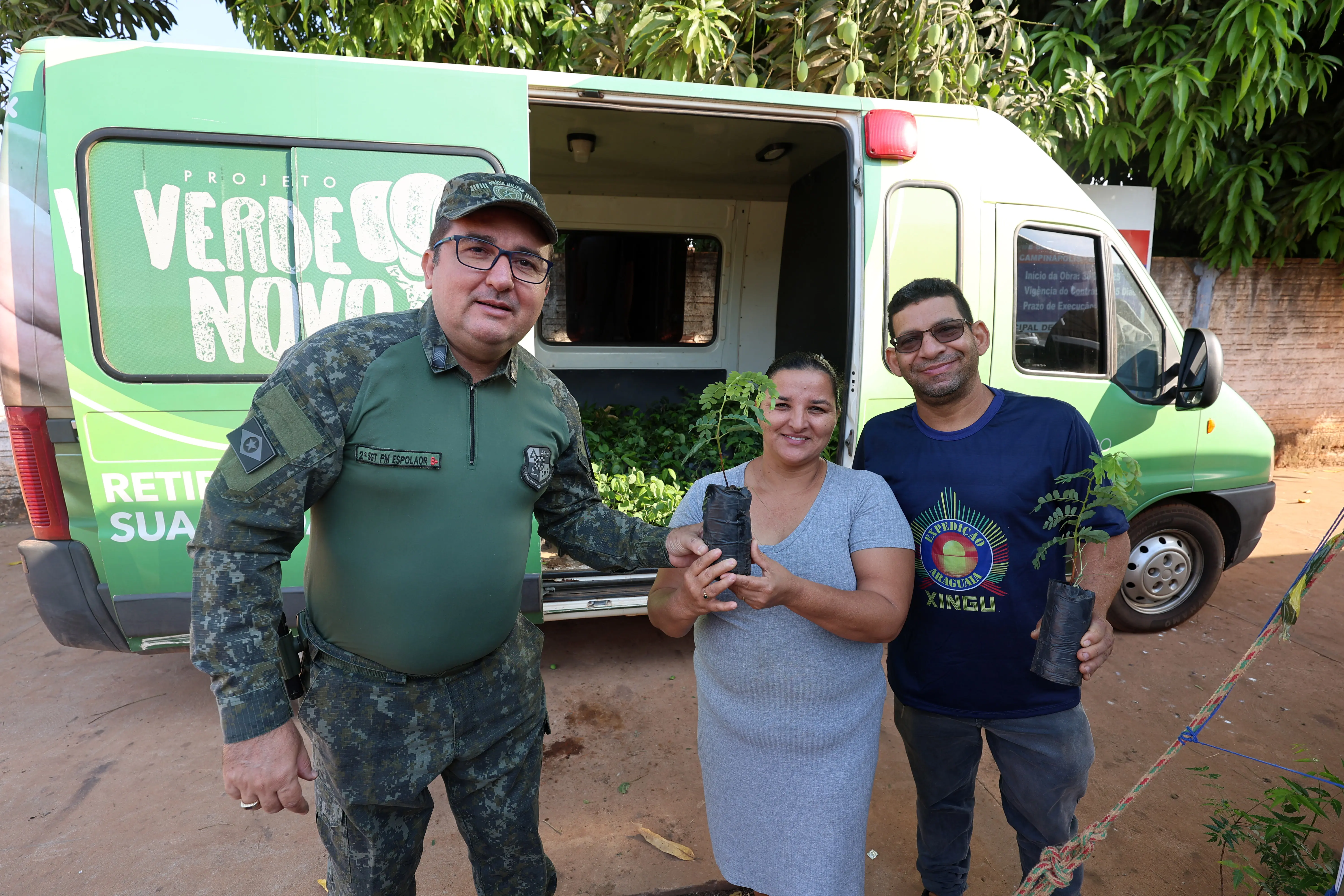 O policial entrega mudas a um casal sorridente diante do veículo do projeto Verde Novo. A ação simboliza parceria entre forças ambientais e comunidade na preservação da natureza