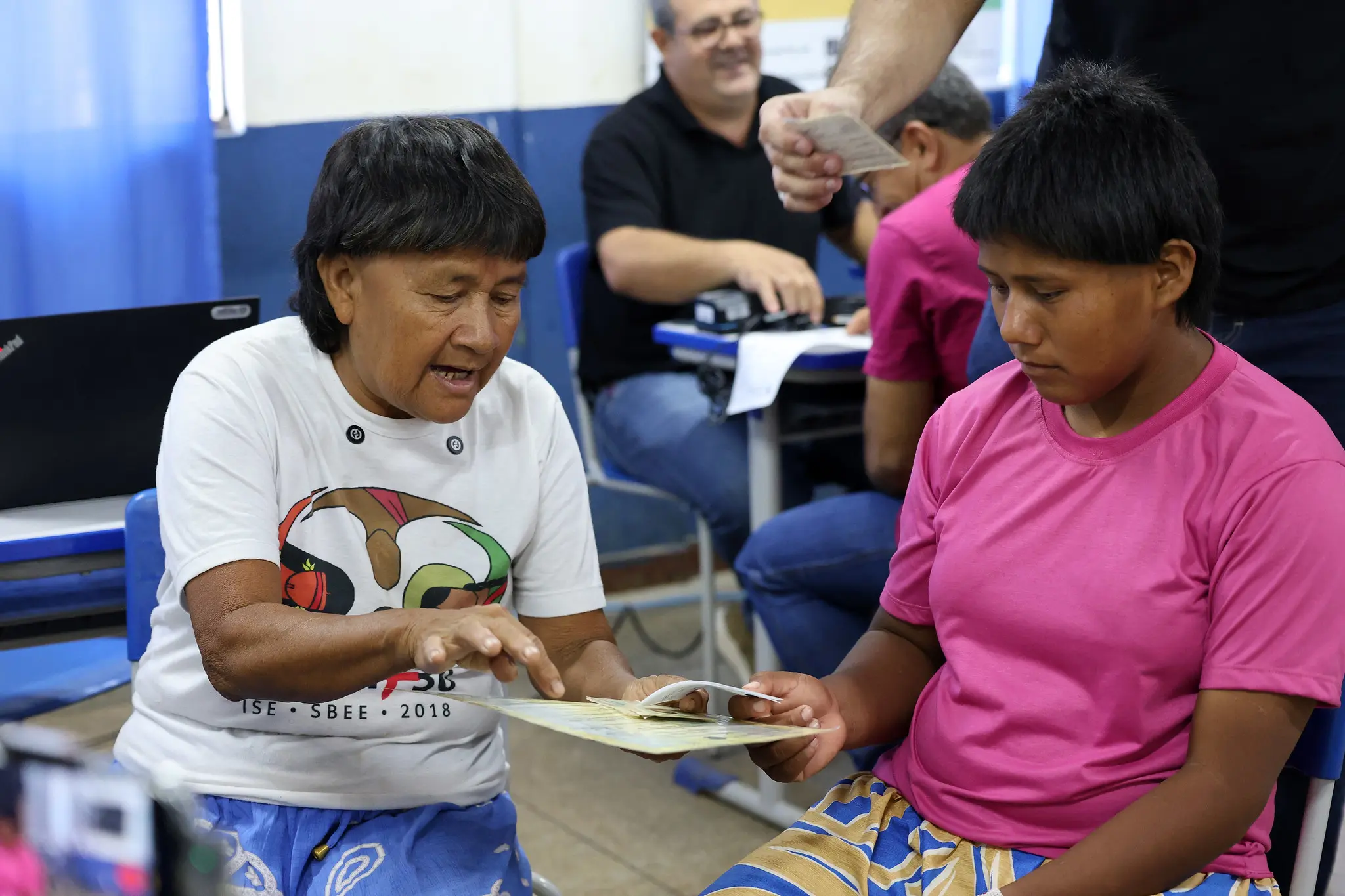 Cacica Carolina e Ana Lúcia. Duas mulheres indígenas de cabelos curtos, com blusas branca e rosa, segurando documentos, durante atendimento em sala do Tribunal Regional Eleitoral de Mato Grosso. 