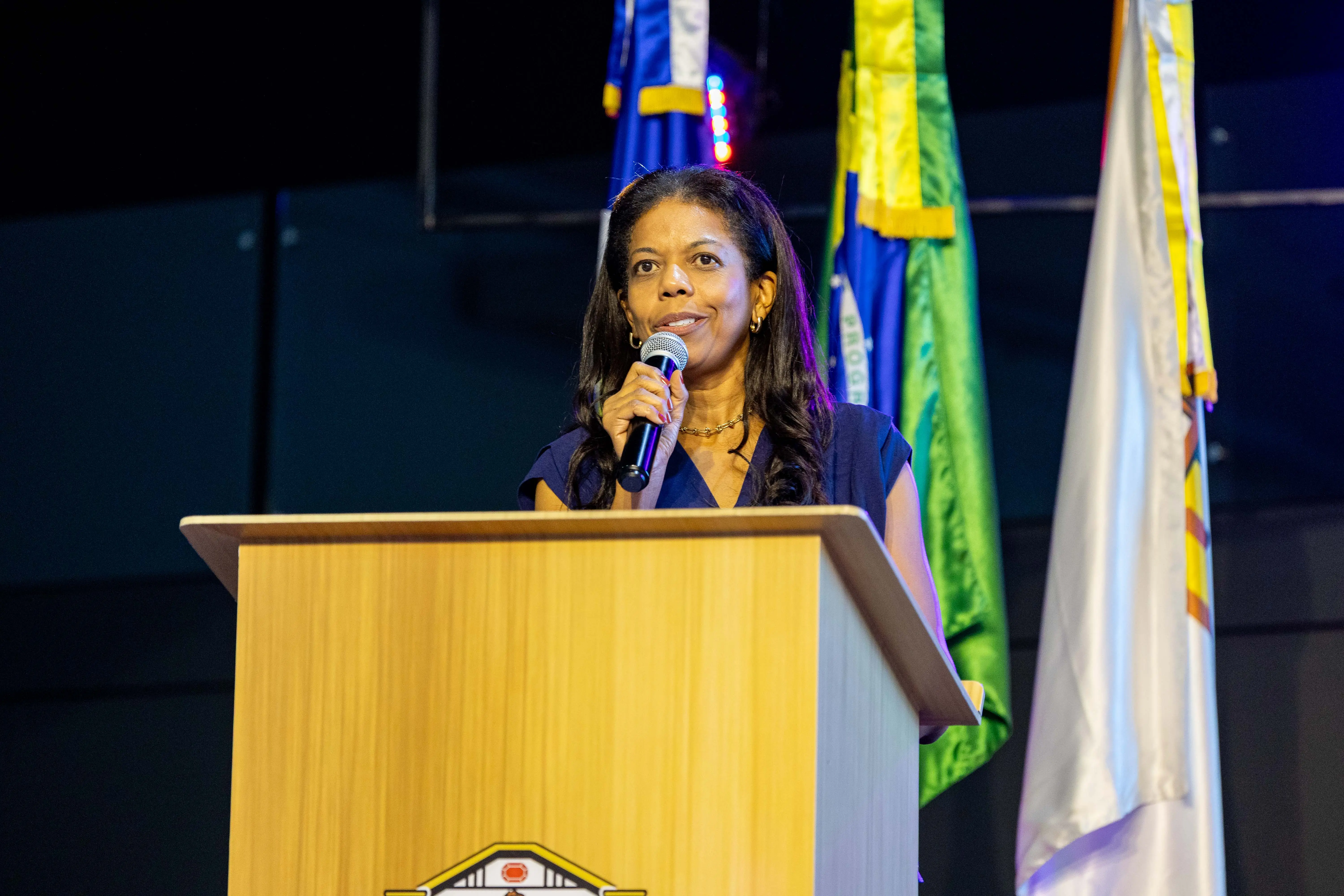 Juíza Renata Evaristo do Carmo fala ao público no auditório do Poder Judiciário de Mato Grosso, em Rondonópolis. Ela está atrás do púlpito com microfone, ao lado das bandeiras do Brasil, Mato Grosso e TJMT.