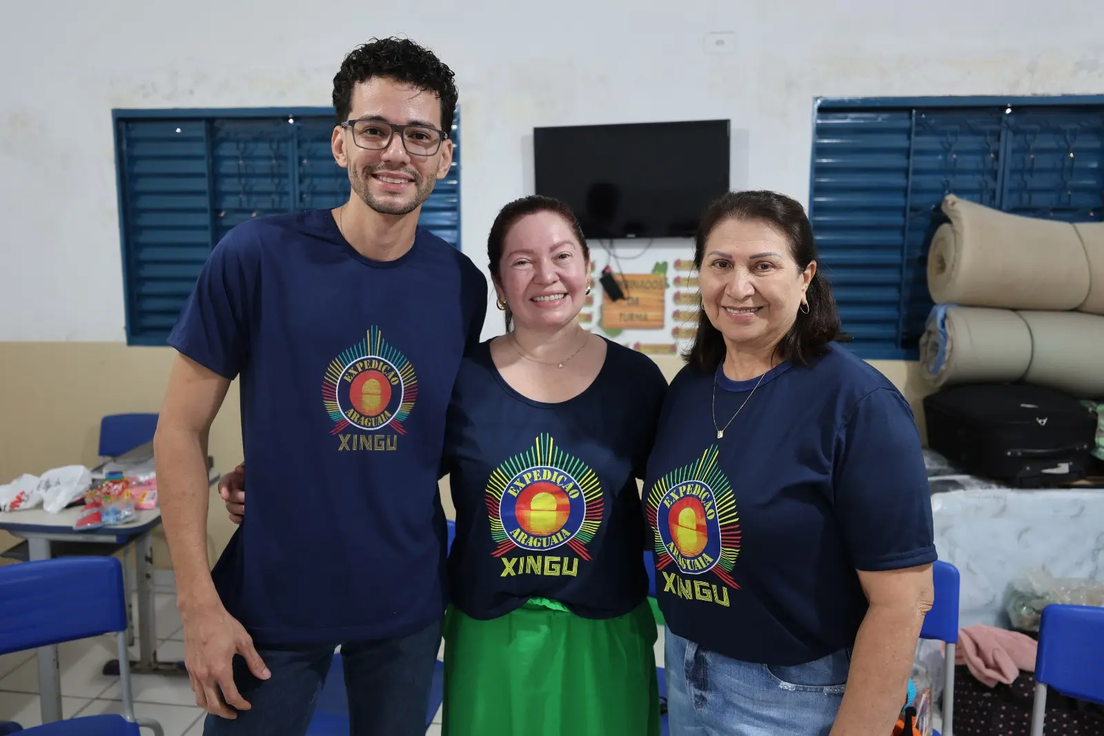 Três integrantes da Expedição Araguaia-Xingu sorriem juntos em sala de aula, vestindo camisetas do projeto. Ao fundo, mesas e materiais de apoio reforçam o clima de trabalho colaborativo