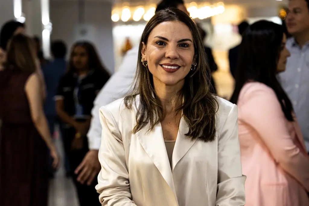A juíza Henriqueta é uma mulher branca, de cabelos castanhos aos ombros, ela está vestindo um blazer branco e uma blusa bege