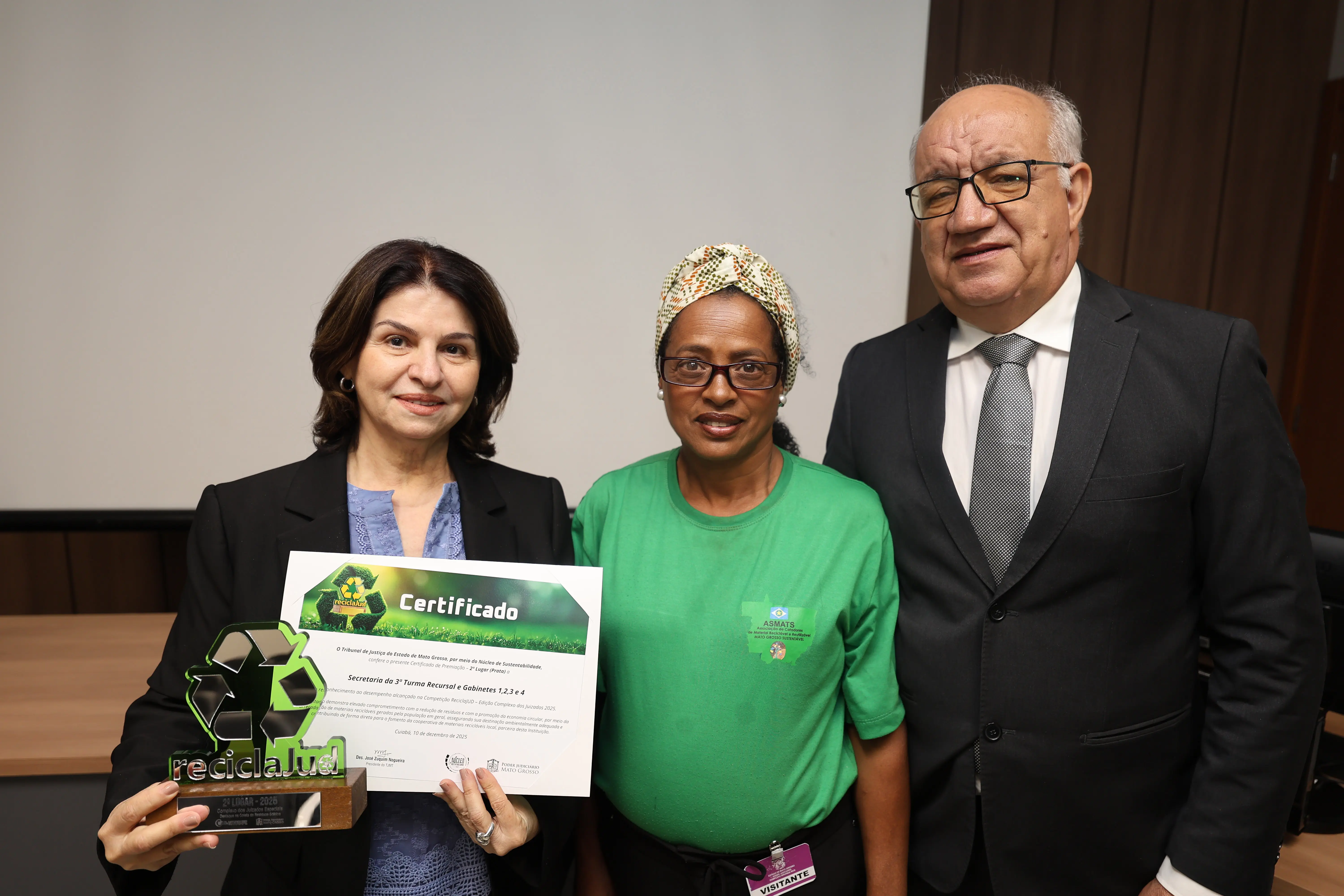 Em sala de reuniões do TJMT, três pessoas posam com certificado e troféu ReciclaJud. À esquerda, magistrada de blazer preto segura a premiação; ao centro, servidora da limpeza com camiseta verde; à direita, homem de terno cinza sorri discretamente. Fundo neutro e ambiente institucional.