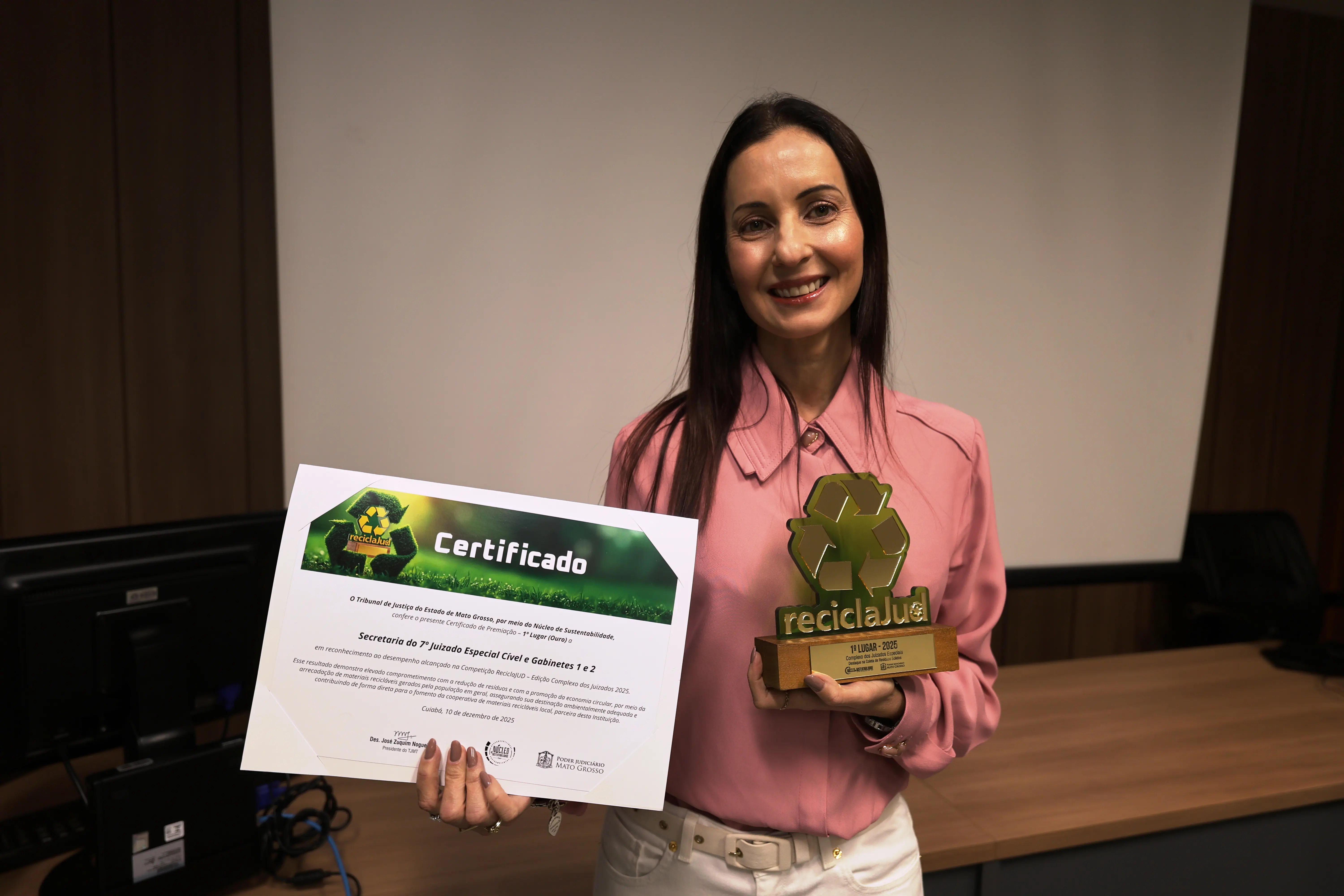 Mulher de camisa rosa e calça clara segura certificado e troféu ReciclaJud em sala de reuniões do TJMT. Ela sorri à frente de painel branco de projeção e mesas com computadores. O ambiente é formal, iluminado e organizado.
