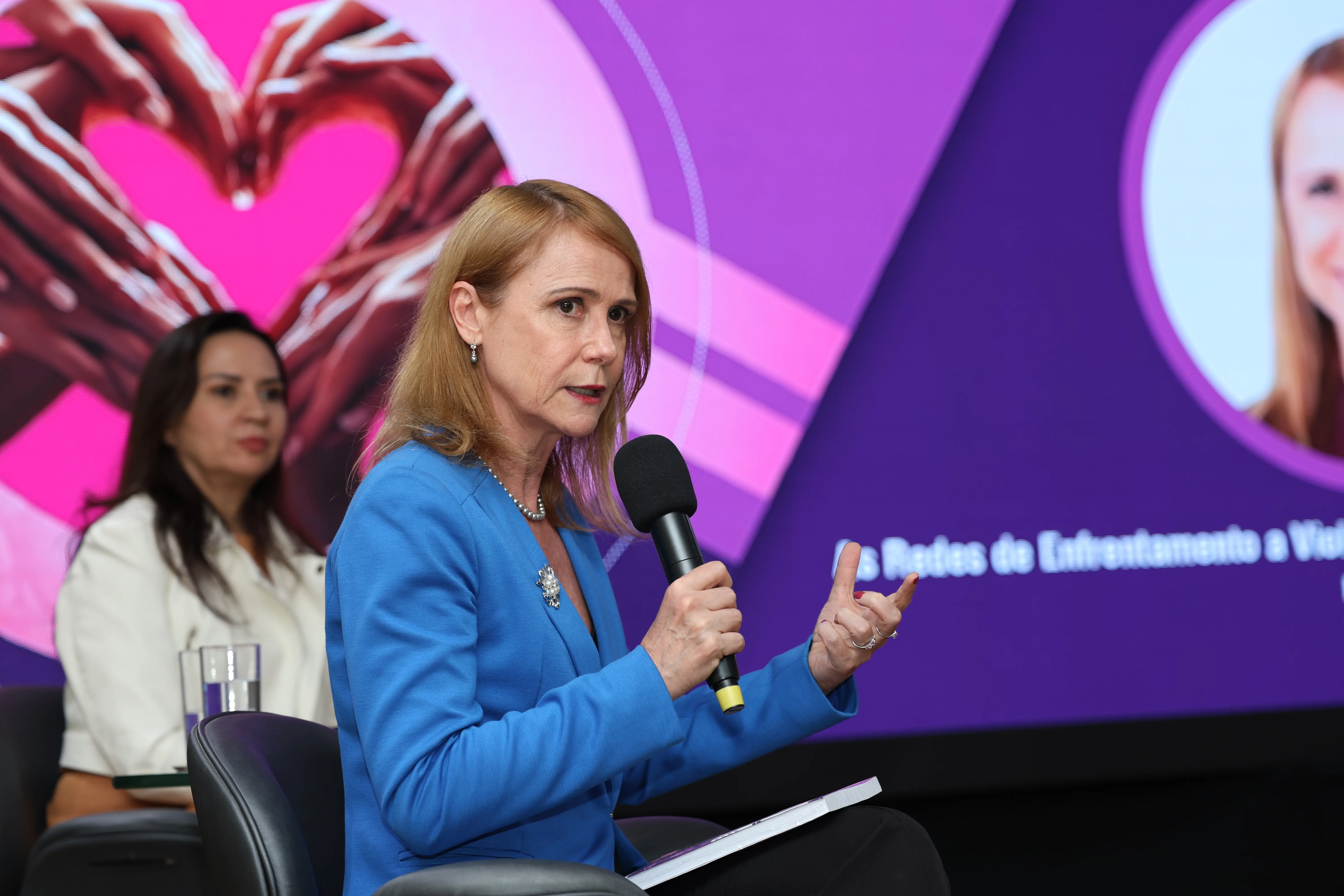 a palestrante Alice Bianchini fala ao microfone durante o II Encontro das Redes de Enfrentamento. Vestindo blazer azul, ela gesticula enquanto explica o tema ao público. Ao fundo, uma integrante das redes acompanha sentada, com o telão colorido do evento