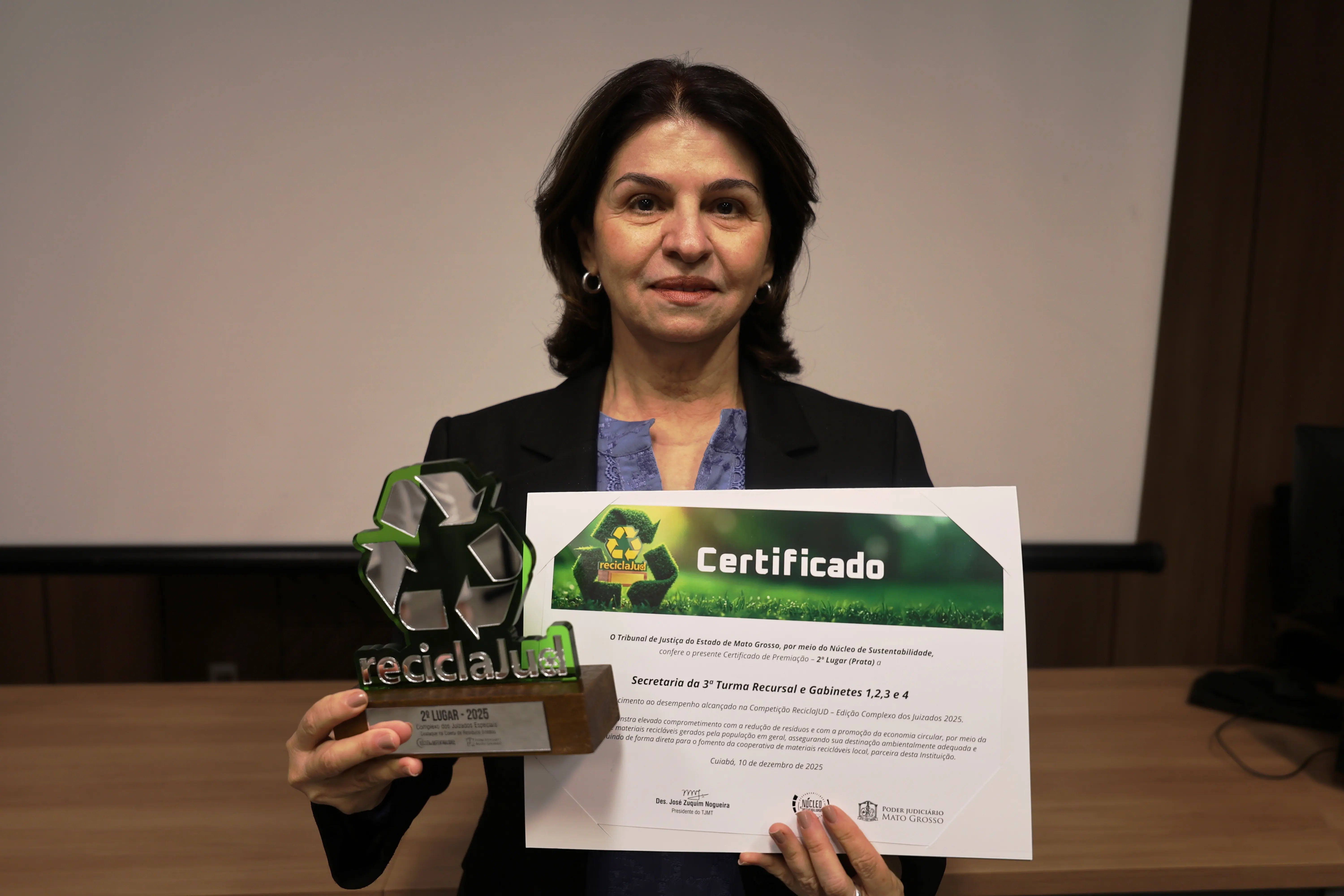 Magistrada de blazer preto segura certificado e troféu ReciclaJud, posando diante de parede branca e mesa institucional. Olhar sério, postura firme. O fundo mostra parte da estrutura da sala de apresentações, com projeção apagada ao fundo.
