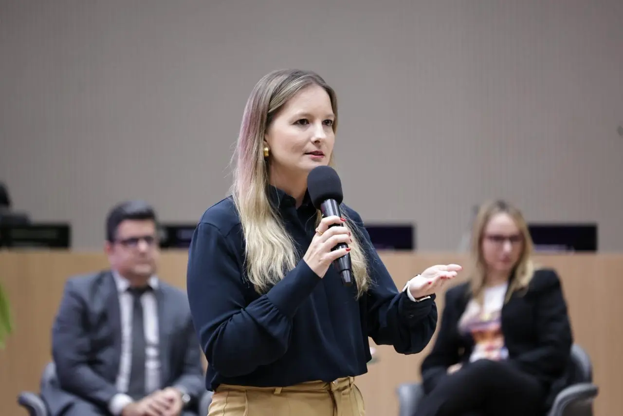 A juíza Djéssika é jovem de cabelos loiros e blusa azul-marinho, fala ao microfone em auditório do TRE. Atrás, duas participantes sentadas acompanham atentas. O ambiente é amplo, com iluminação suave e mesas de apoio ao fundo.