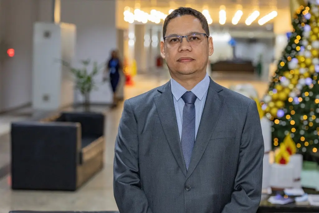 O juiz Luís Felipe é um homem branco, de cabelos escuros curto e óculos. Ele usa um terno cinza claro, camisa azul listrada e uma gravata azul.