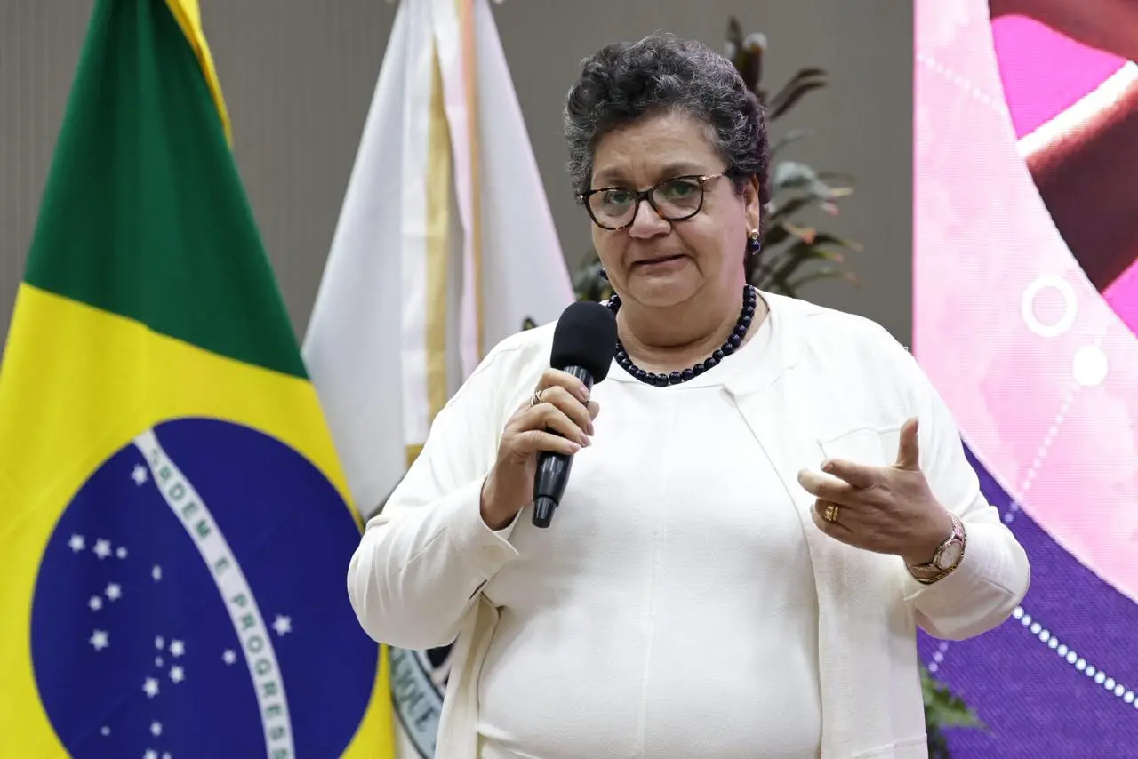 A expositora, juíza Maria Mazarello é uma senhora de cabelos curtos e óculos, veste conjunto claro e colar azul. Fala ao microfone diante das bandeiras do Brasil e de MT. O auditório do TRE aparece ao fundo com painel iluminado em rosa.