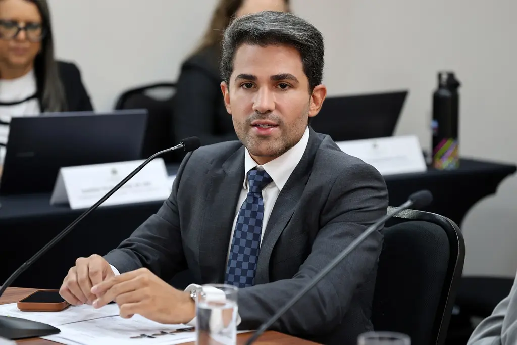 Foto horizontal que mostra o coordenador do Projeto Conecta, desembargador Pedro Felipe de Oliveira Santos, sentado, falando ao microfone. Ele é um homem jovem, da olhos castanhos, cabelos grisalhos, usando camisa branca, terno cinza e gravata xadrez em tons de azul.