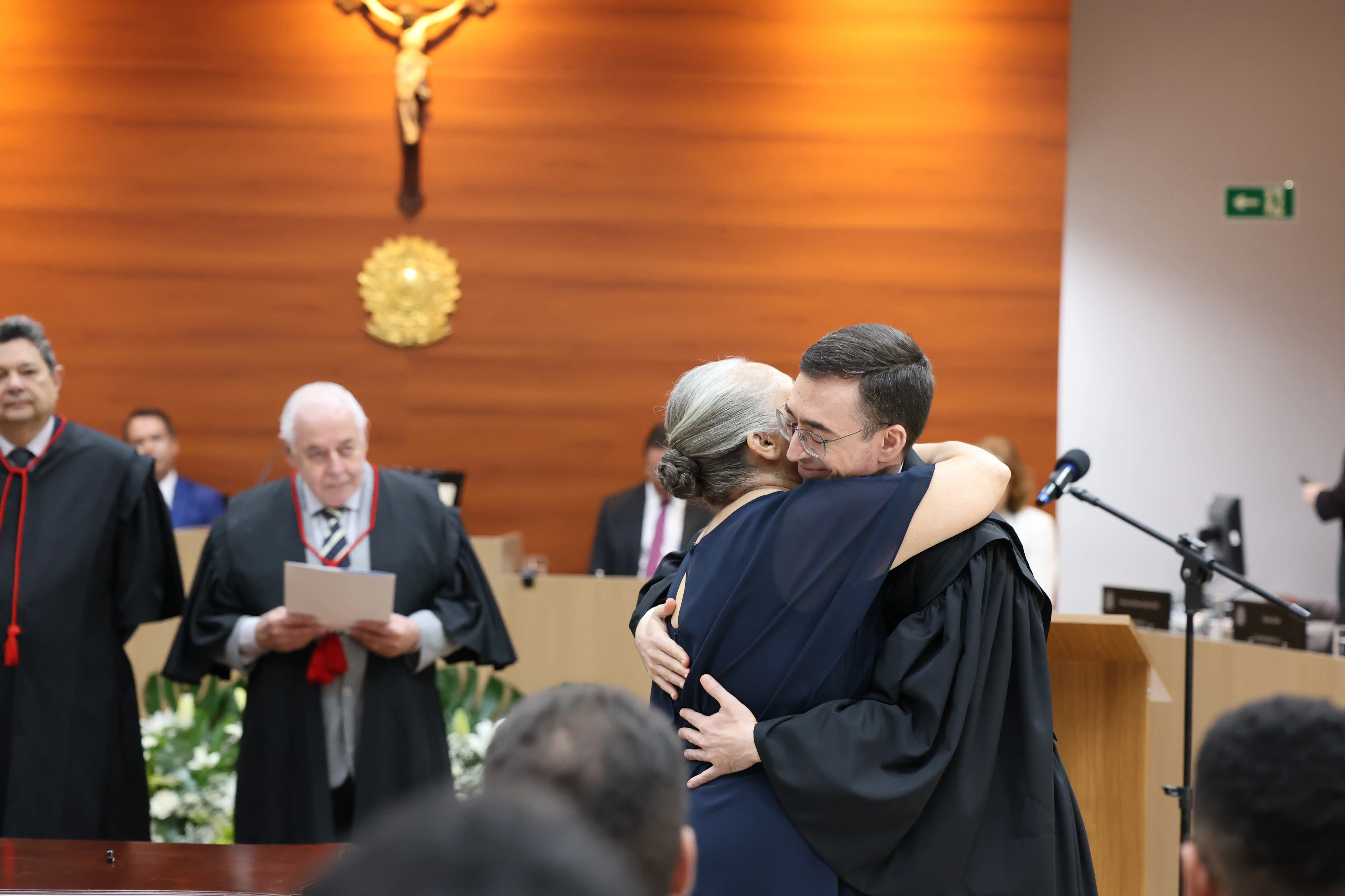 Magistrado de toga recebe o colar simbólico durante cerimônia de posse, em gesto afetuoso com familiar. A cena ocorre no plenário do TJMT, com autoridades e parede de madeira ao fundo.