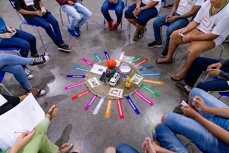 Foto horizontal que mostra vários estudantes adolescentes participando de um círculo de construção de paz. Ele estão sentados em círculo em volta de objetos colocados no chão, como papéis coloridos com palavras impressas, brinquedos, entre outros.