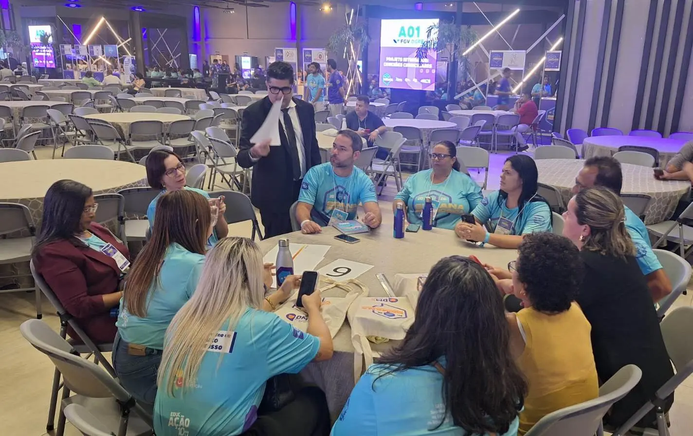 Foto horizontal ambientada em um grande salão de eventos. Em primeiro plano, professores usando a camiseta da Semana Pedagógica estão sentados em volta de uma mesa redonda. Em pé, o juiz Marcos Terêncio os orienta sobre a oficina sobre violência doméstica. 