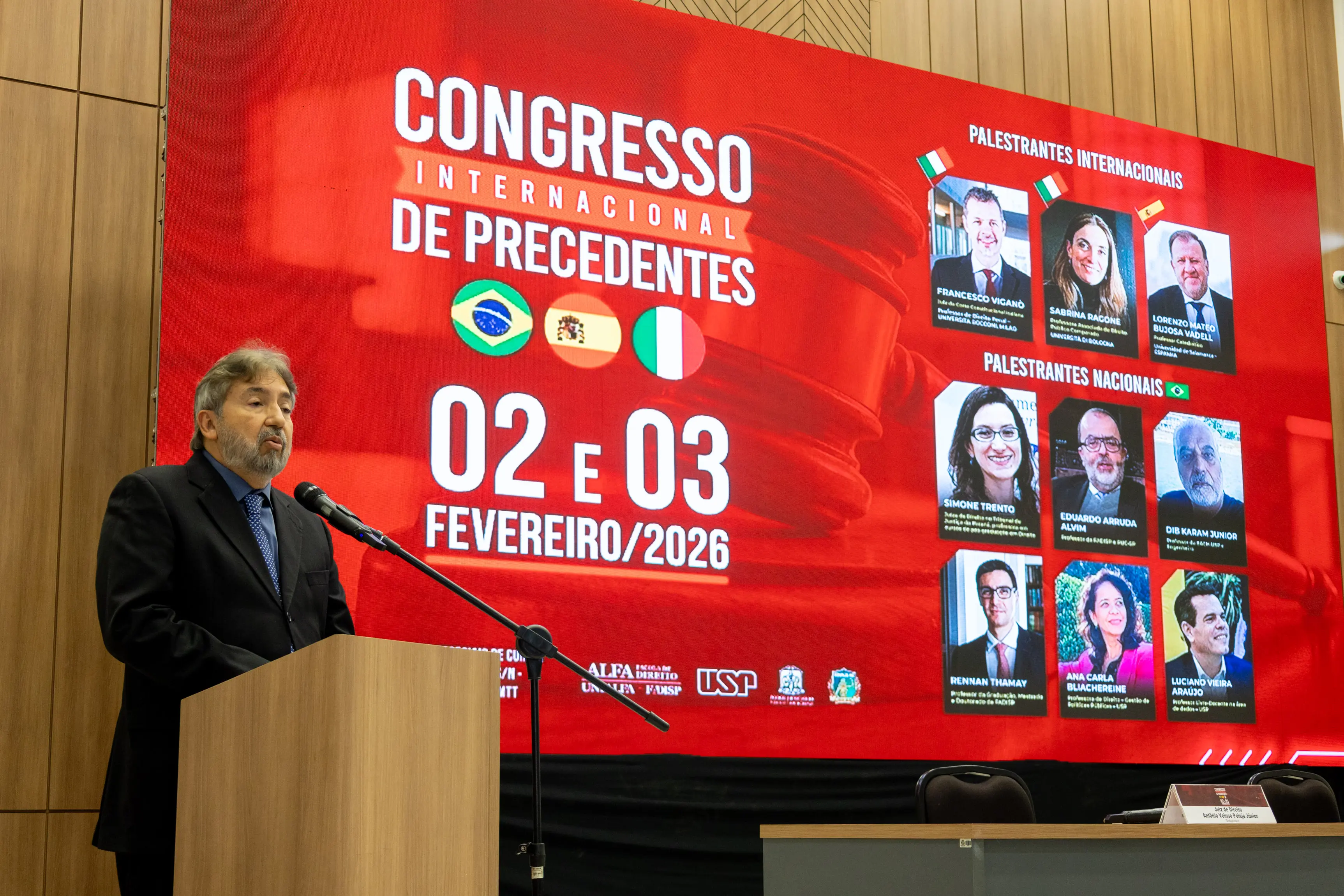 Um homem de barba e terno discursa em um púlpito. Ao fundo, um grande telão vermelho exibe "CONGRESSO INTERNACIONAL DE PRECEDENTES" com fotos de palestrantes e as datas 02 e 03 de fevereiro de 2026.