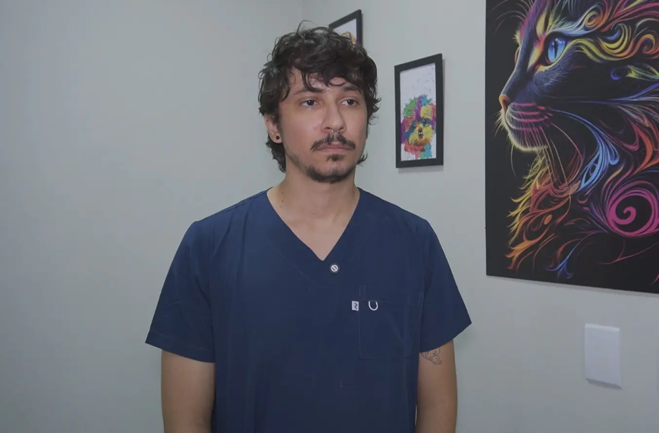 Foto horizontal em plano médio que mostra o médico veterinário João Meireles em pé, da cintura pra cima. Ele é um jovem de pele parda, olhos, cabelos e barba escuros, usando jaleco azul marinho. Na parede do consultório dele, há quadros com ilustrações de gatos e cachorros. 
