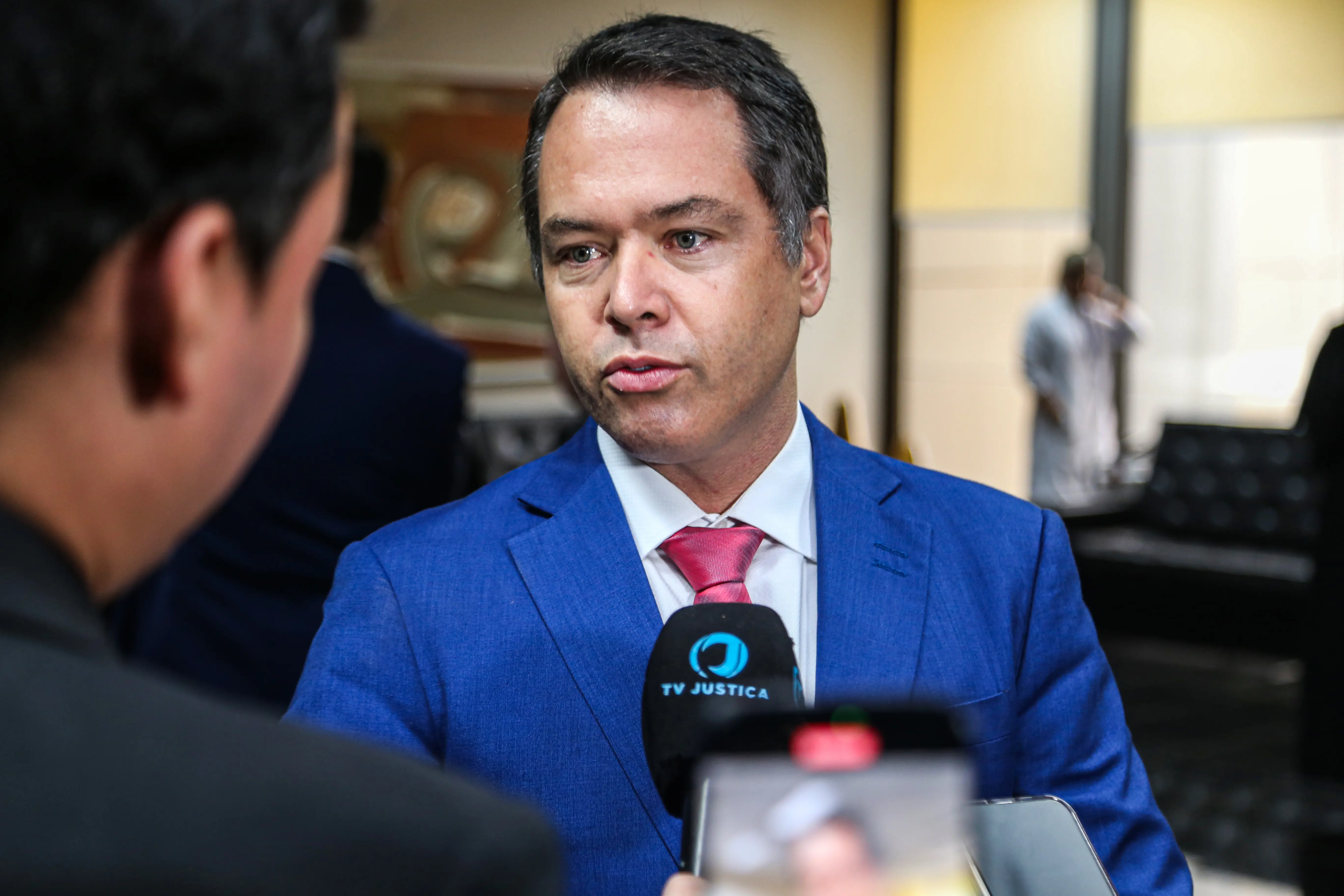 Homem de terno azul fala à TV Justiça. Microfone à frente, ambiente corporativo ao fundo. Gravata rosa destaca no traje formal.