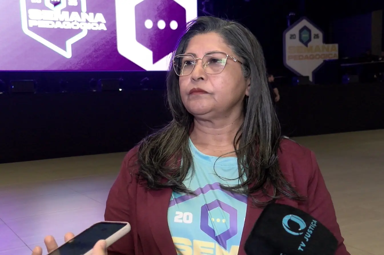 Foto horizontal que mostra a professora F�tima Ramos durante entrevista � TV Justi�a. Ela � uma mulher parda, de cabelos longos, lisos e grisalhos, usando camiseta azul clara da Semana Pedag�gica, blazer vinho e �culos de grau. Atr�s dela est� o palco com a logo da Semana Pedag�gica 2026.