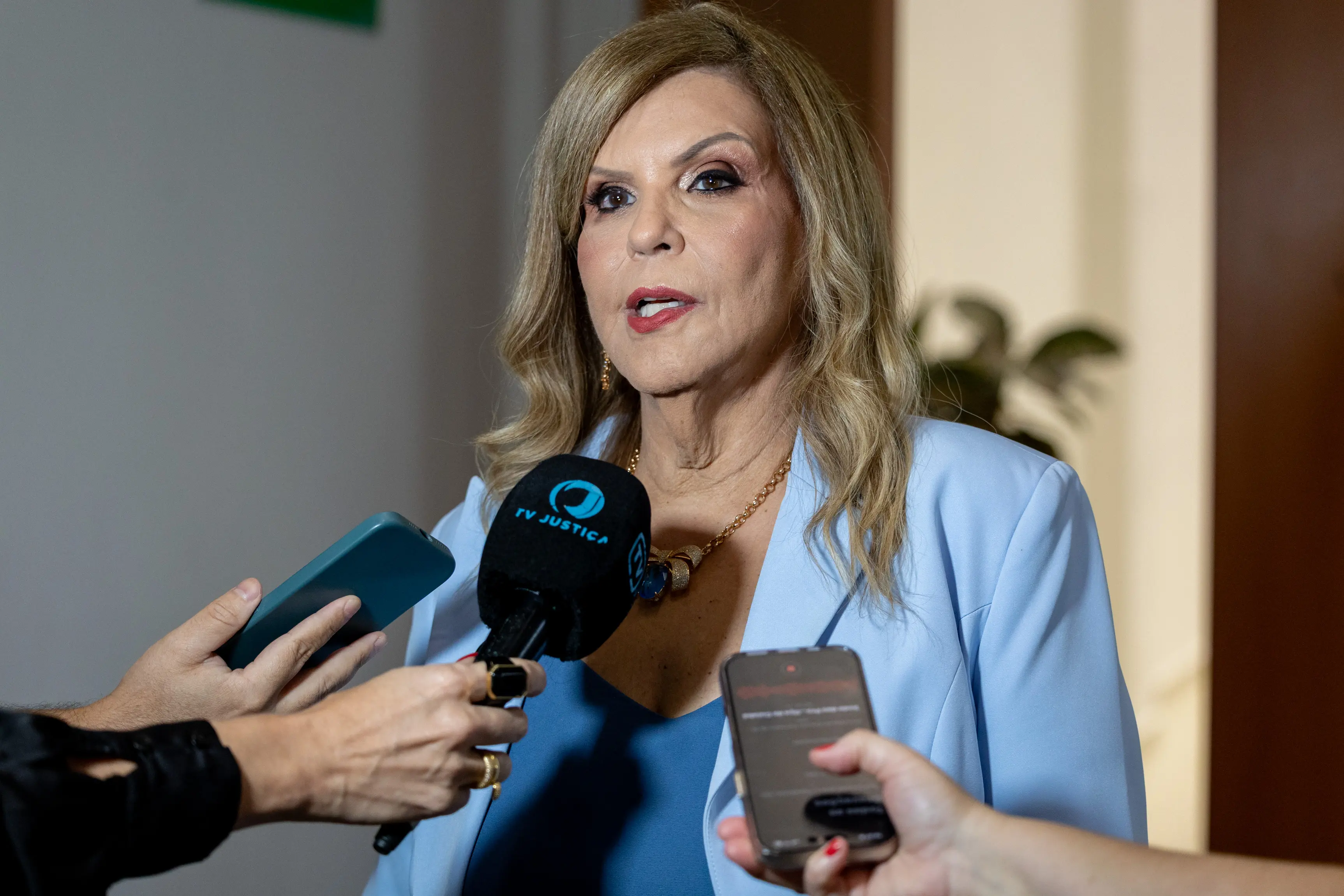 Uma mulher loira de blazer azul claro e blusa azul-marinho concede entrevista. Ela usa um colar dourado e fala em direção a um microfone da "TV JUS" e a um celular que registra a conversa.