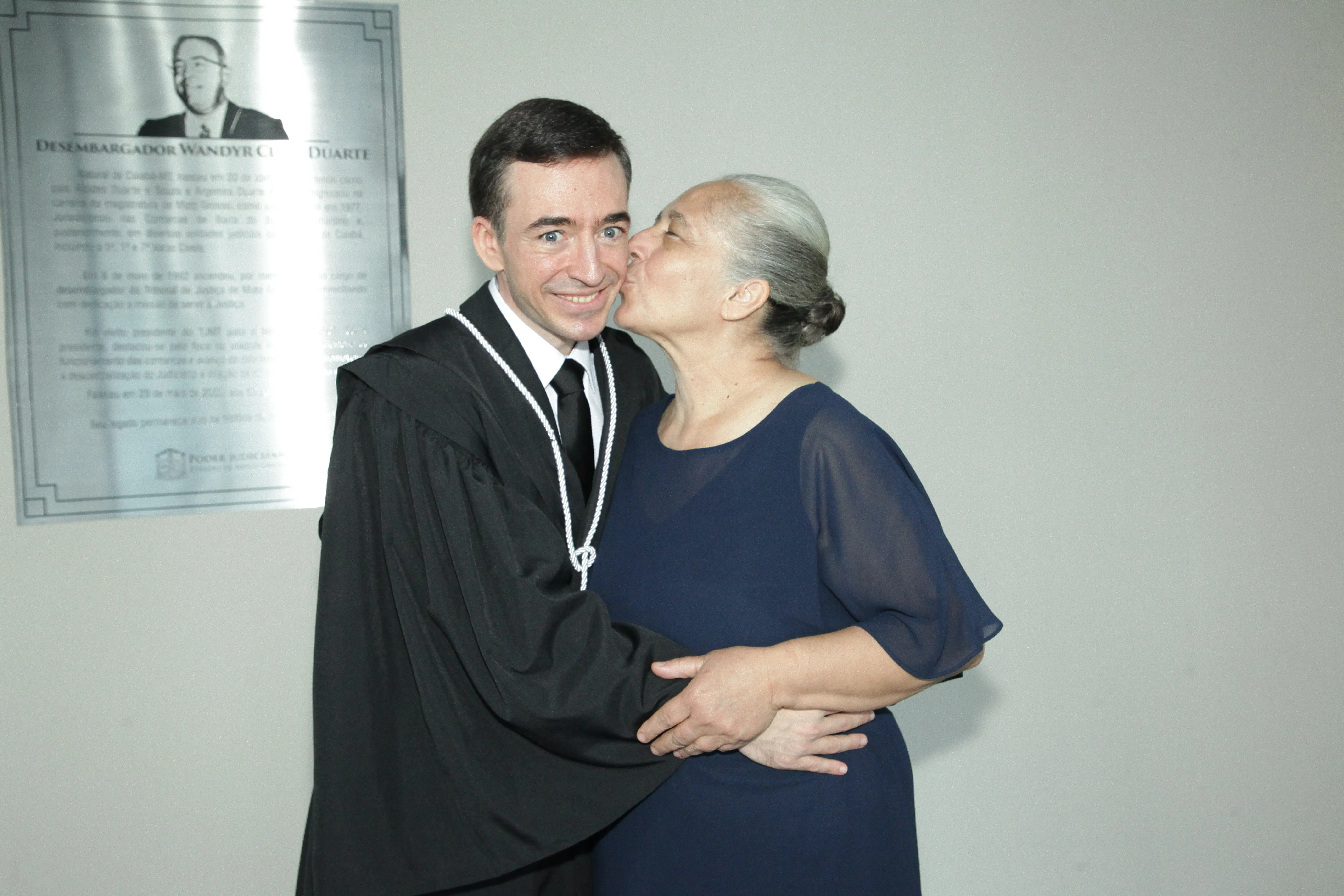 Magistrado de toga posa ao lado da mãe, em gesto afetuoso. A imagem foi registrada em espaço interno do Tribunal, com paredes claras e clima de solenidade.