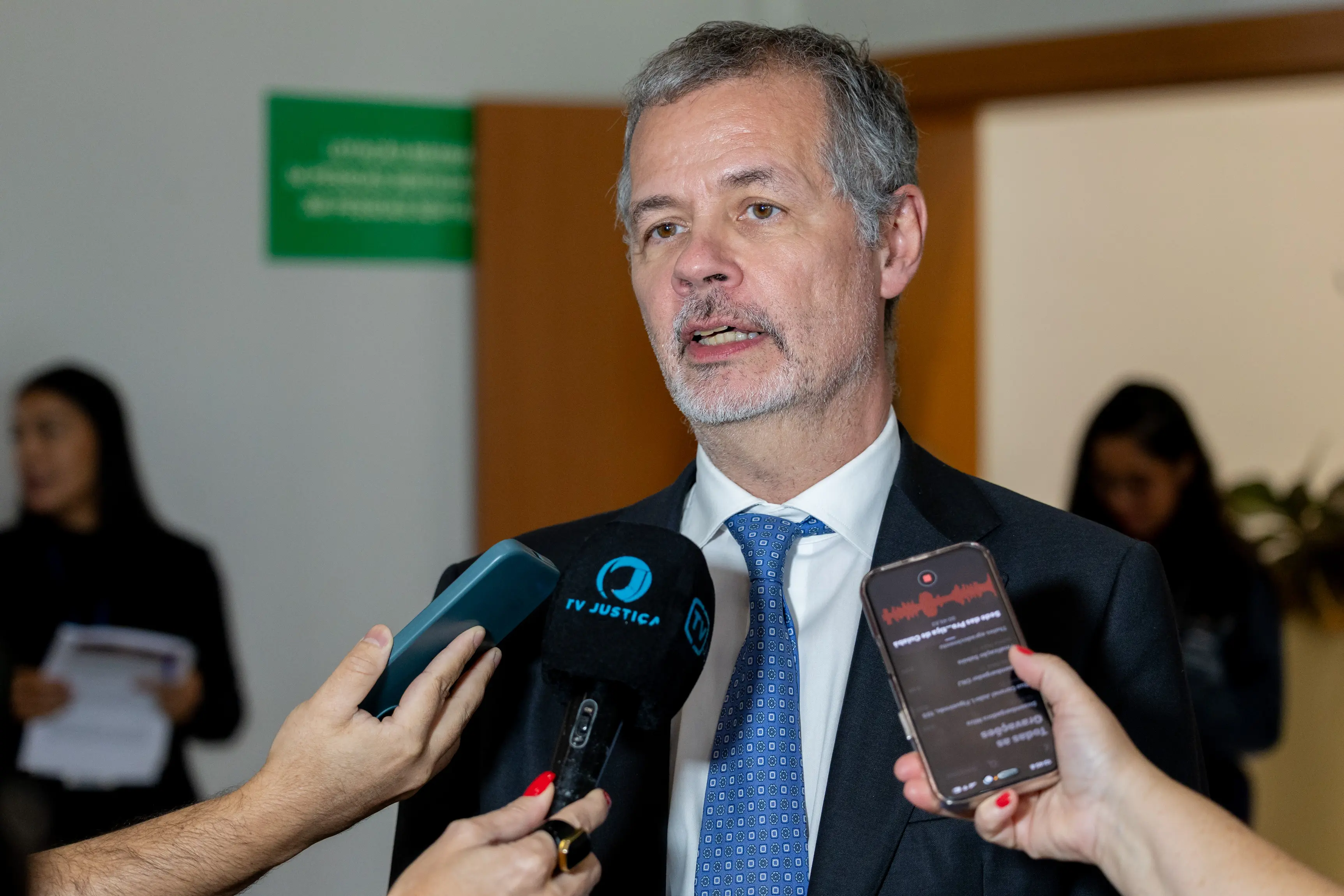 Um homem de pele clara e cabelos grisalhos curtos, vestindo terno e gravata azul, concede entrevista. Ele fala para um microfone da "TV JUSTIÇA" e dois celulares que gravam sua fala.