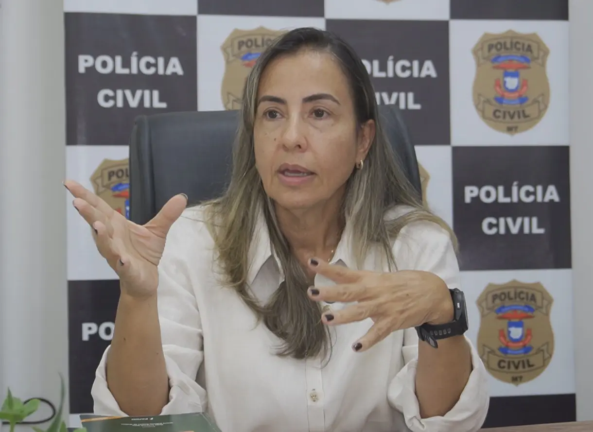 foto horizontal que mostra a delegada de polícia Liliane Murata sentada em seu escritório e gesticulando, durante uma entrevista. Ela é uma mulher de pele clara, cabelos longos lisos e com mechas loiras, usando camisa de linho bege. Atrás dela, há um painel com a logomarca da Polícia Civil.