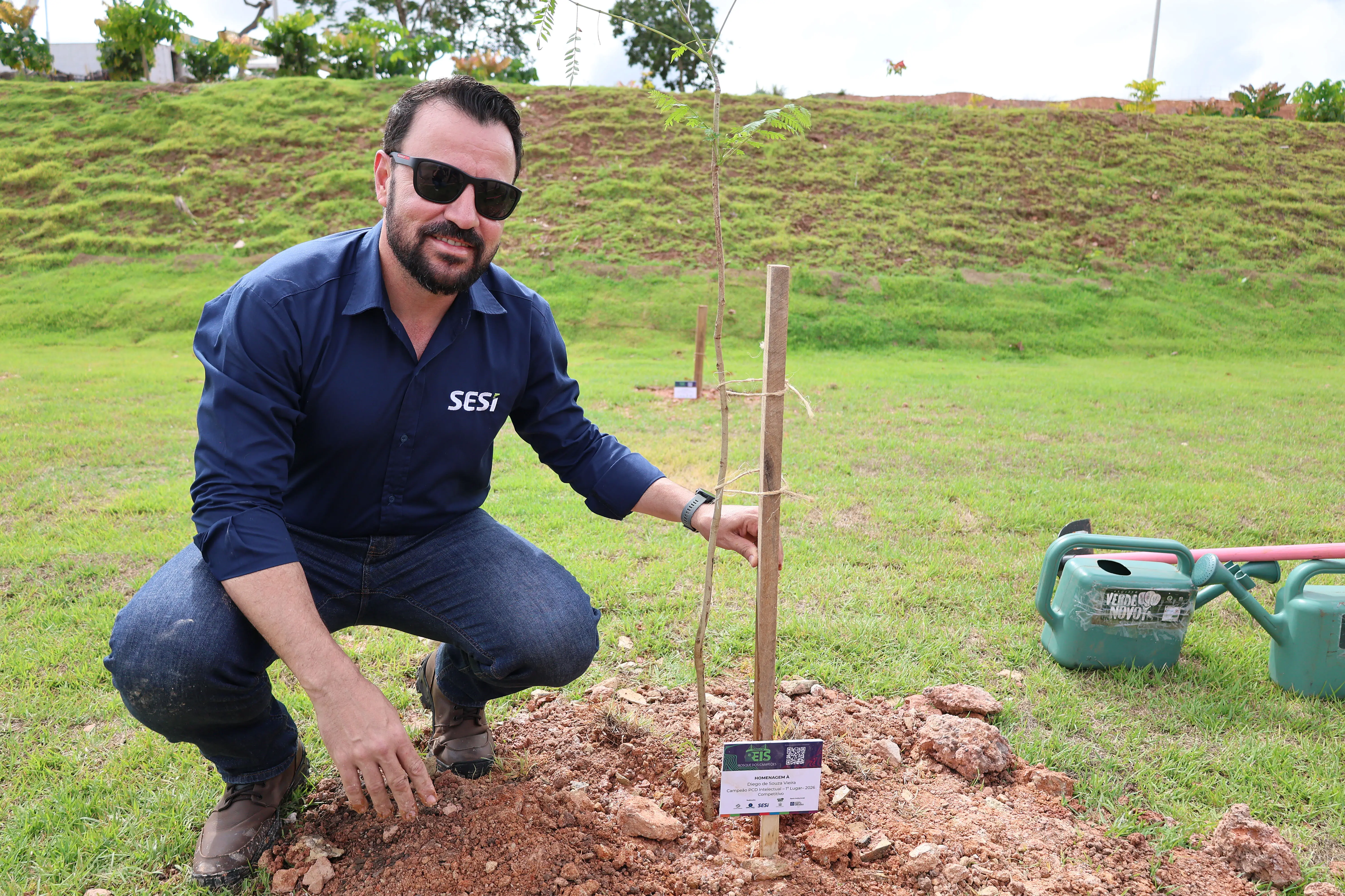  Homem de pele clara, barba escura e óculos de sol, agachado ao lado de uma muda de árvore recém-plantada. Veste camisa azul-marinho com o logo 