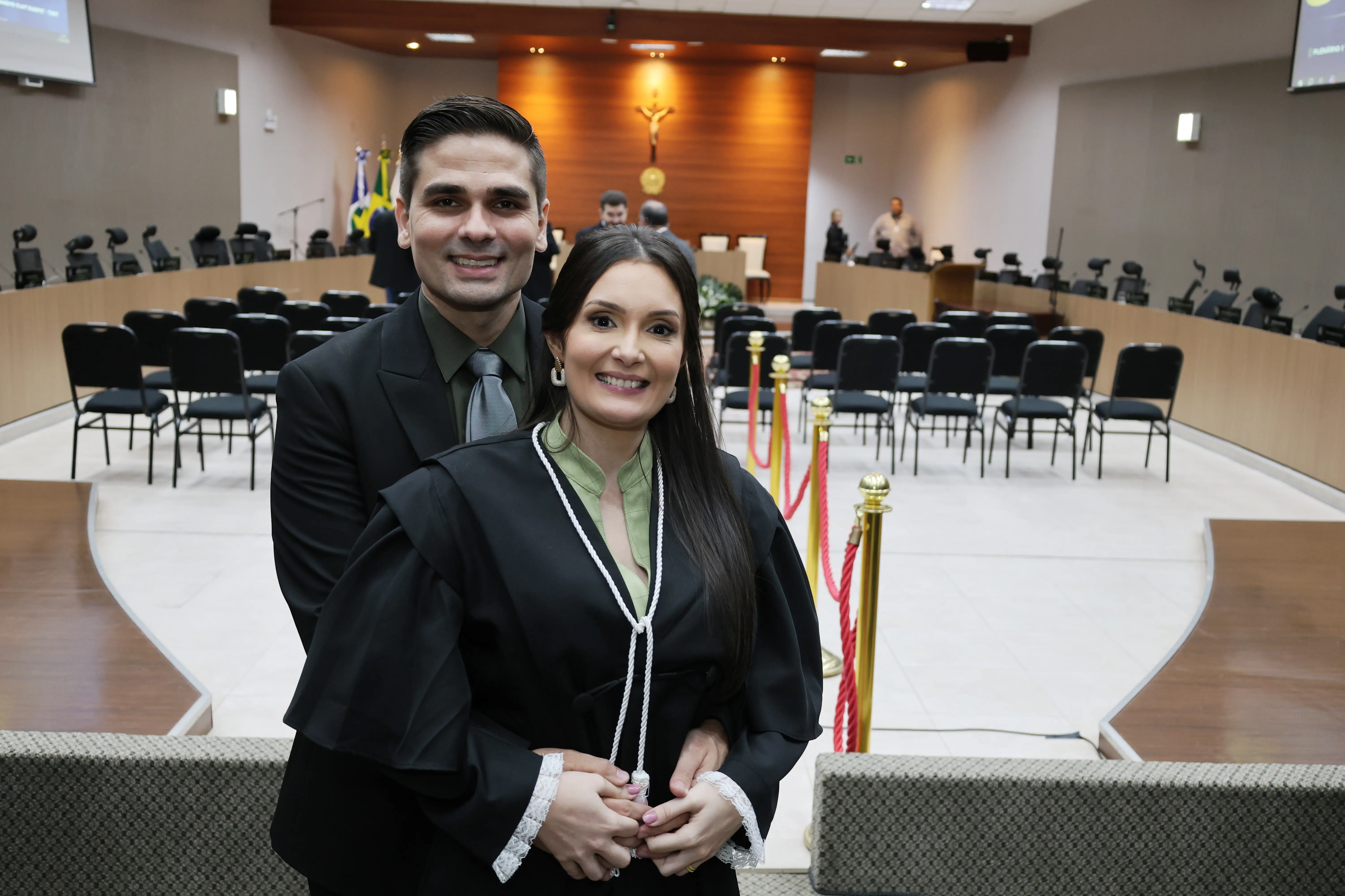 Magistrada posa ao lado do esposo no plenário do TJMT. Ambos estão sorrindo, com cadeiras do colegiado e bandeiras oficiais ao fundo, em ambiente institucional.