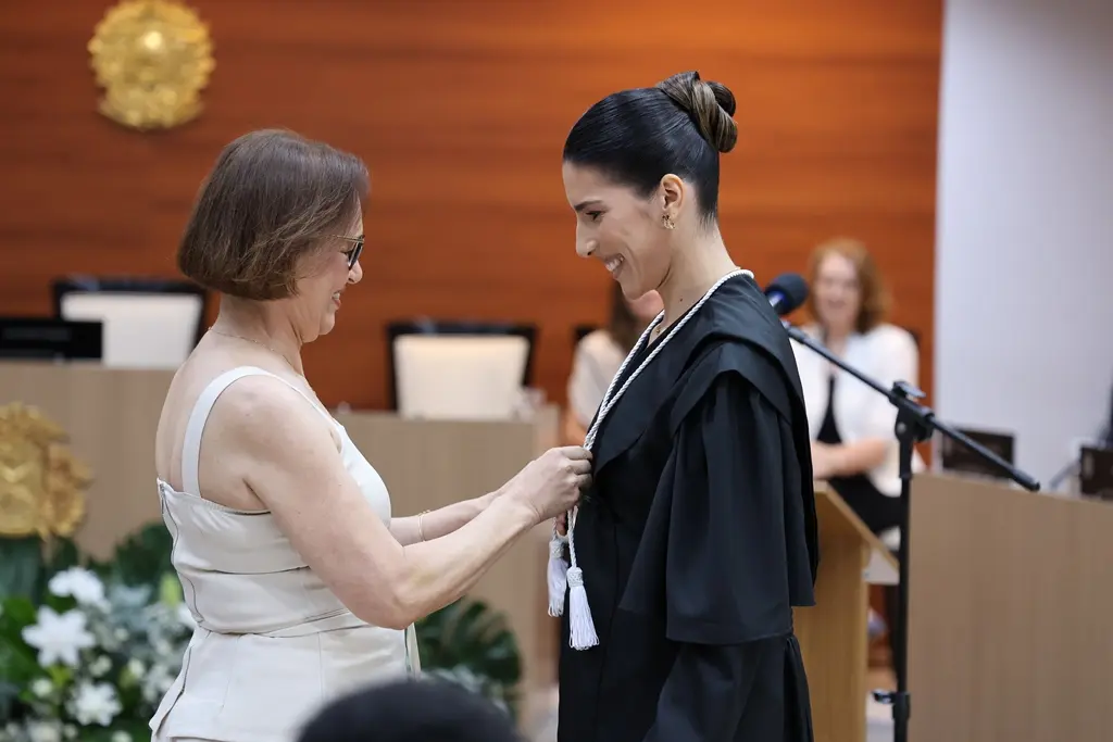 Foto horizontal que mostra a juíza Laís Paranhos de frente para sua mãe, que arruma sua toga, durante sessão de posse. Elas sorriem uma para a outra.