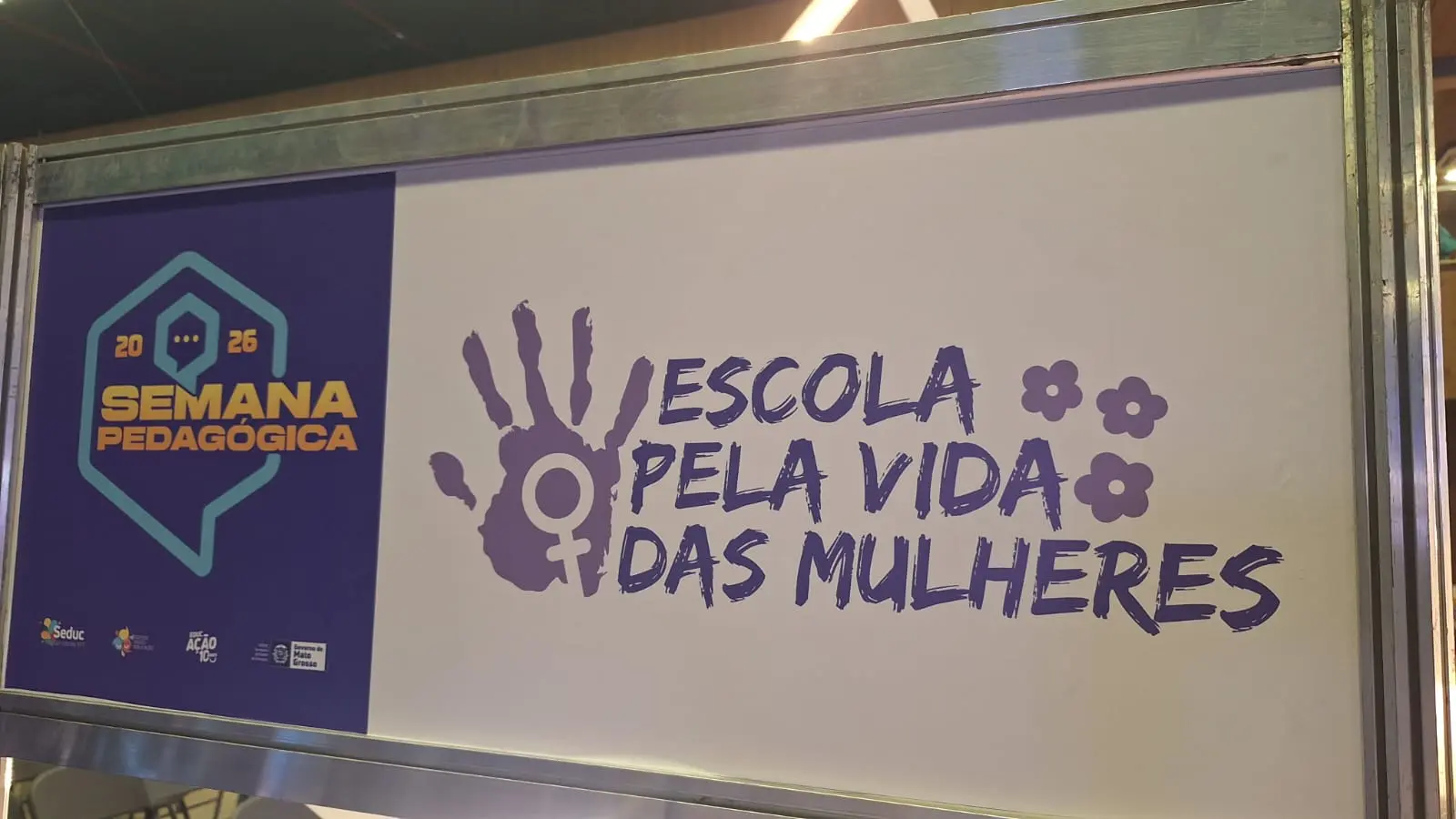 Foto vertical que mostra detalhe do estande da Semana Pedagógica 2026 com o tema “Escola pela vida das mulheres”. Essa frase aparece escrita em letra lilás, com o símbolo de uma mão espalmada com o símbolo do espelho de vênus sobreposto. 