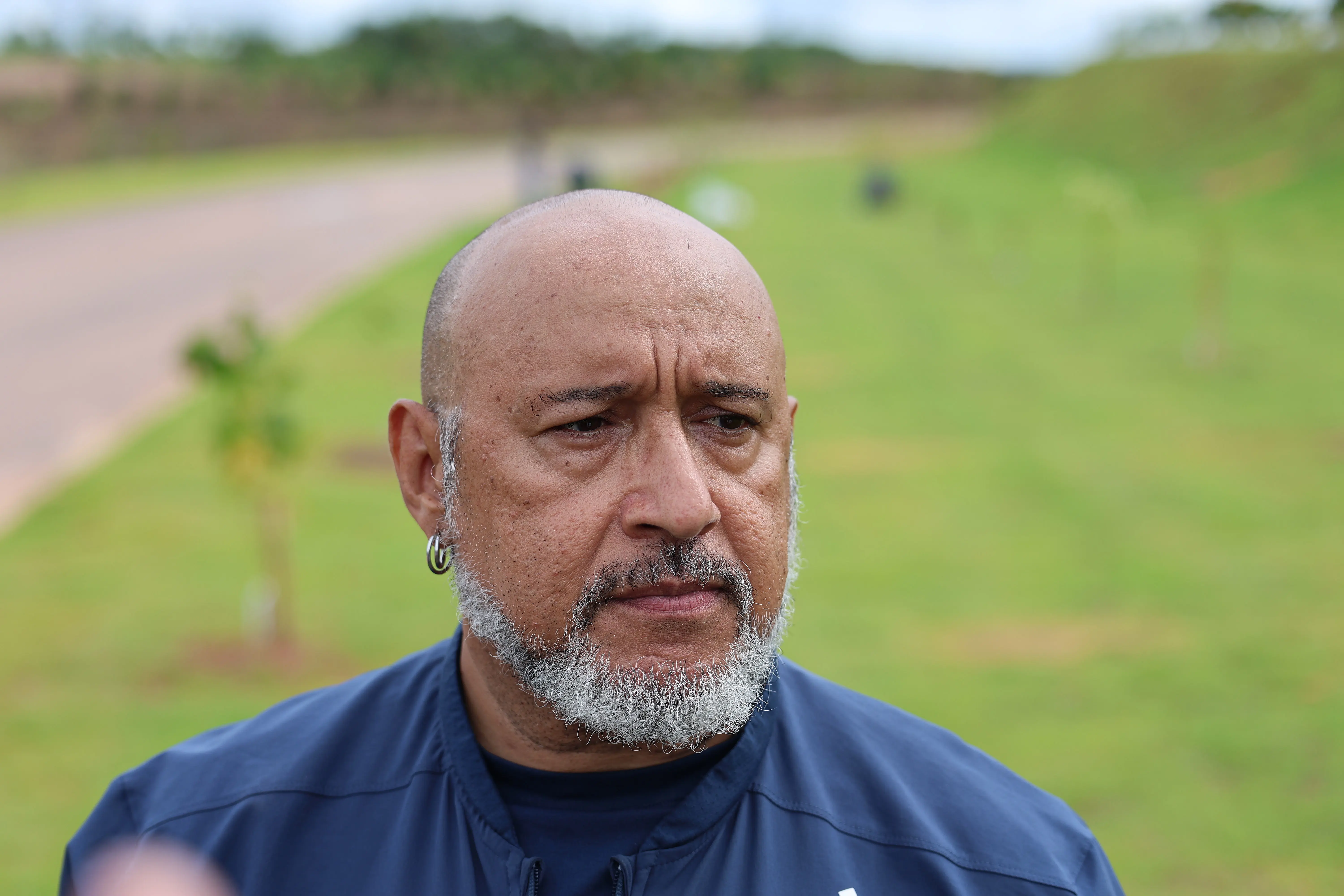 Homem com barba grisalha, de expressão séria. Ele usa uma jaqueta azul-marinho e um brinco de argola na orelha direita. O fundo mostra um campo gramado com pequenas mudas de árvores sob um céu nublado.