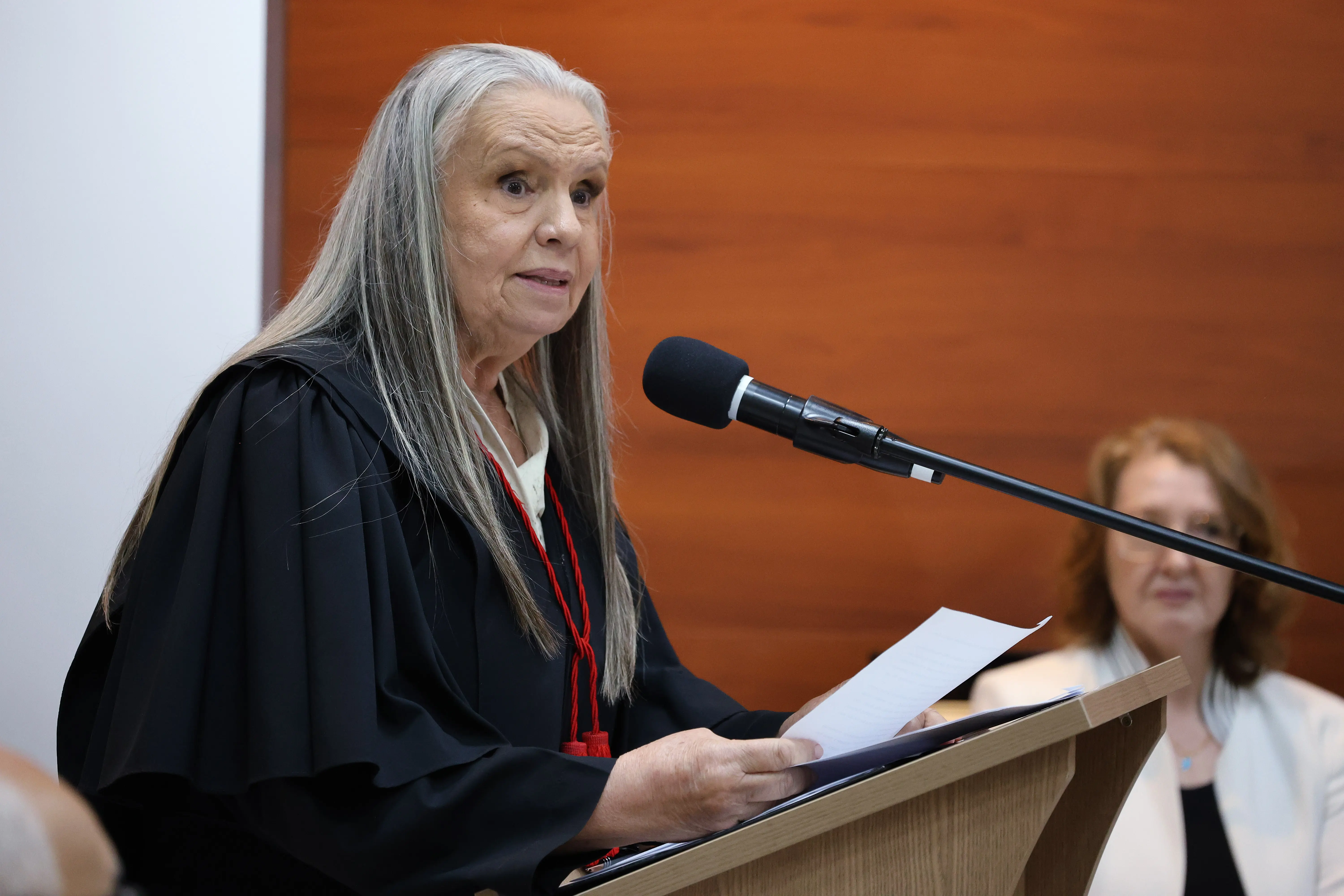 Uma mulher de longos cabelos grisalhos e toga preta lê um documento em um púlpito de madeira. Ela fala ao microfone. Ao fundo, uma mulher de blazer branco observa.