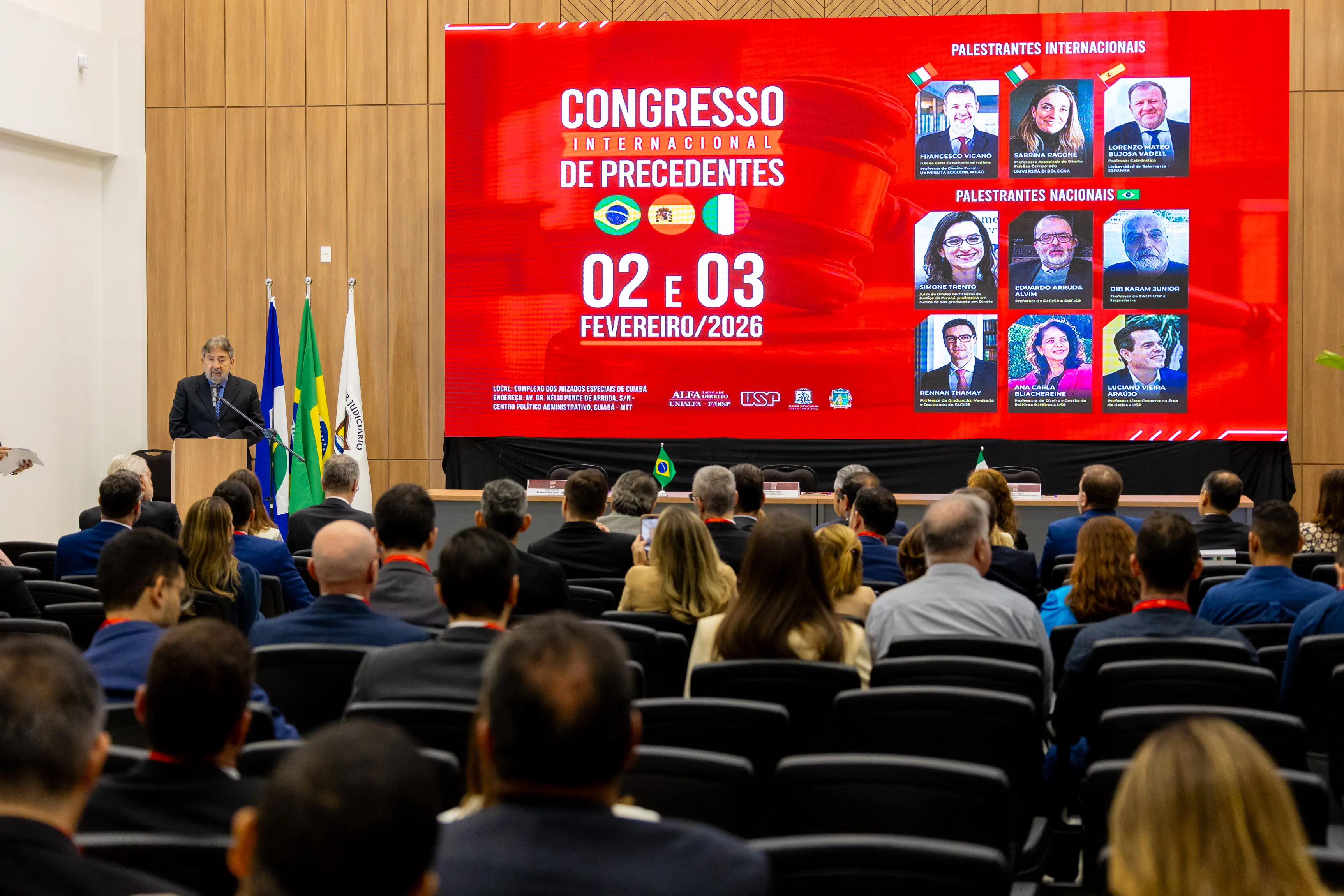 Auditório lotado visto de trás. À frente, um homem discursa em um púlpito ao lado de bandeiras. Ao centro, um grande telão vermelho exibe "CONGRESSO INTERNACIONAL DE PRECEDENTES" e fotos de palestrantes.