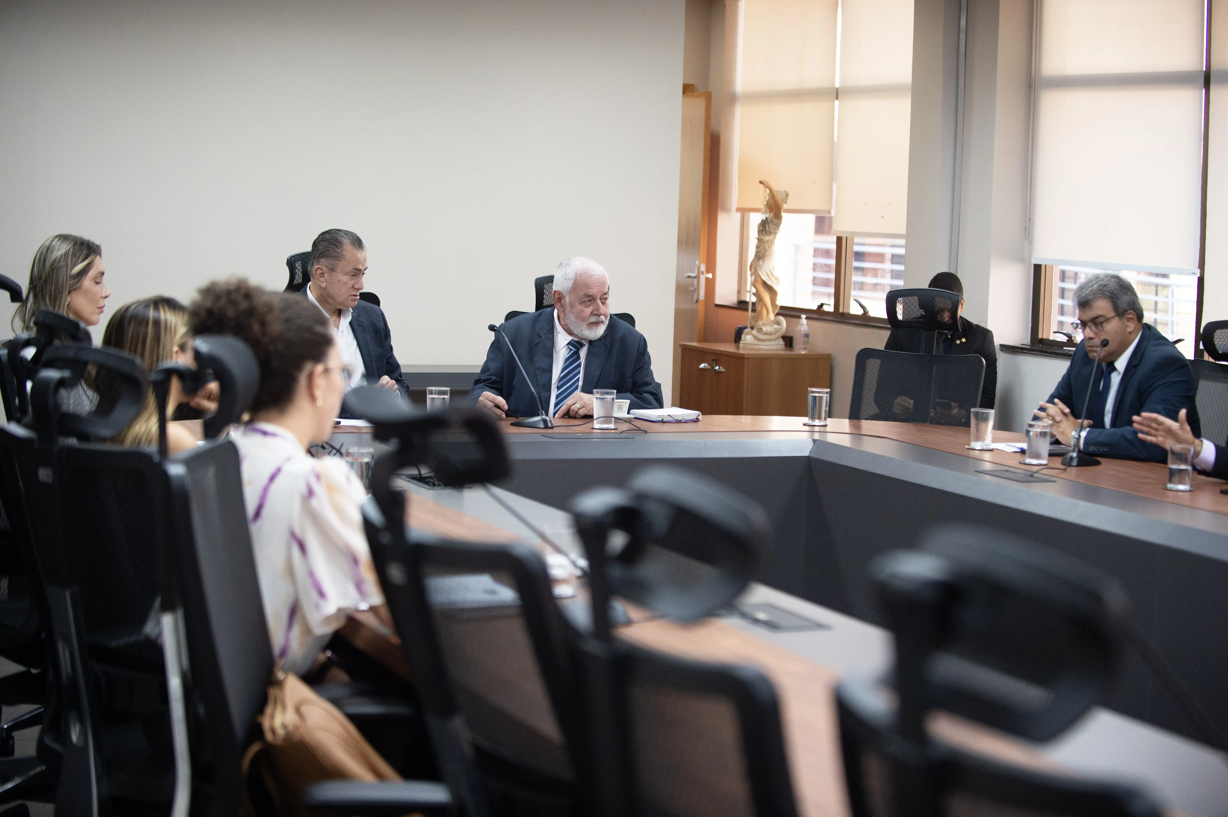 a imagem mostra a sala de reuniões. As nove pessoas estão sentadas à mesa, em formato de U, olhando para o presidente do TJTM, enquanto ele fala. 