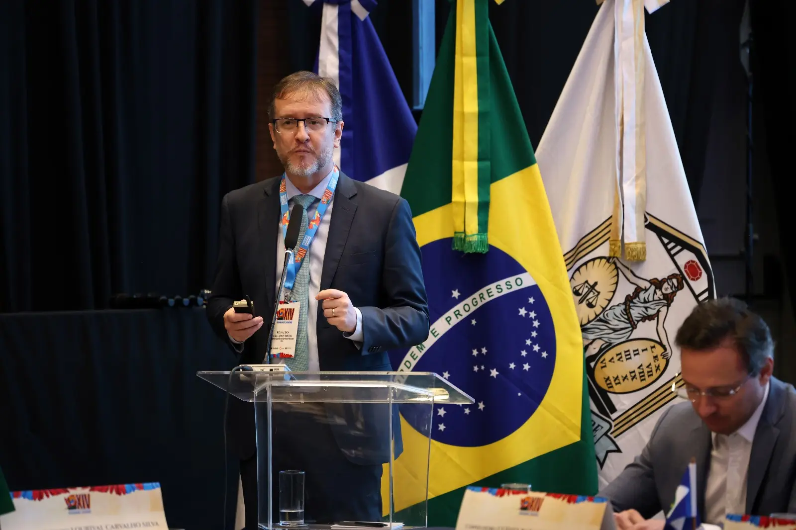 Homem de terno e crachá fala ao microfone em púlpito transparente. Ao fundo, bandeiras do Brasil e institucional reforçam o ambiente formal. Ele segura um controle, indicando apresentação em andamento.