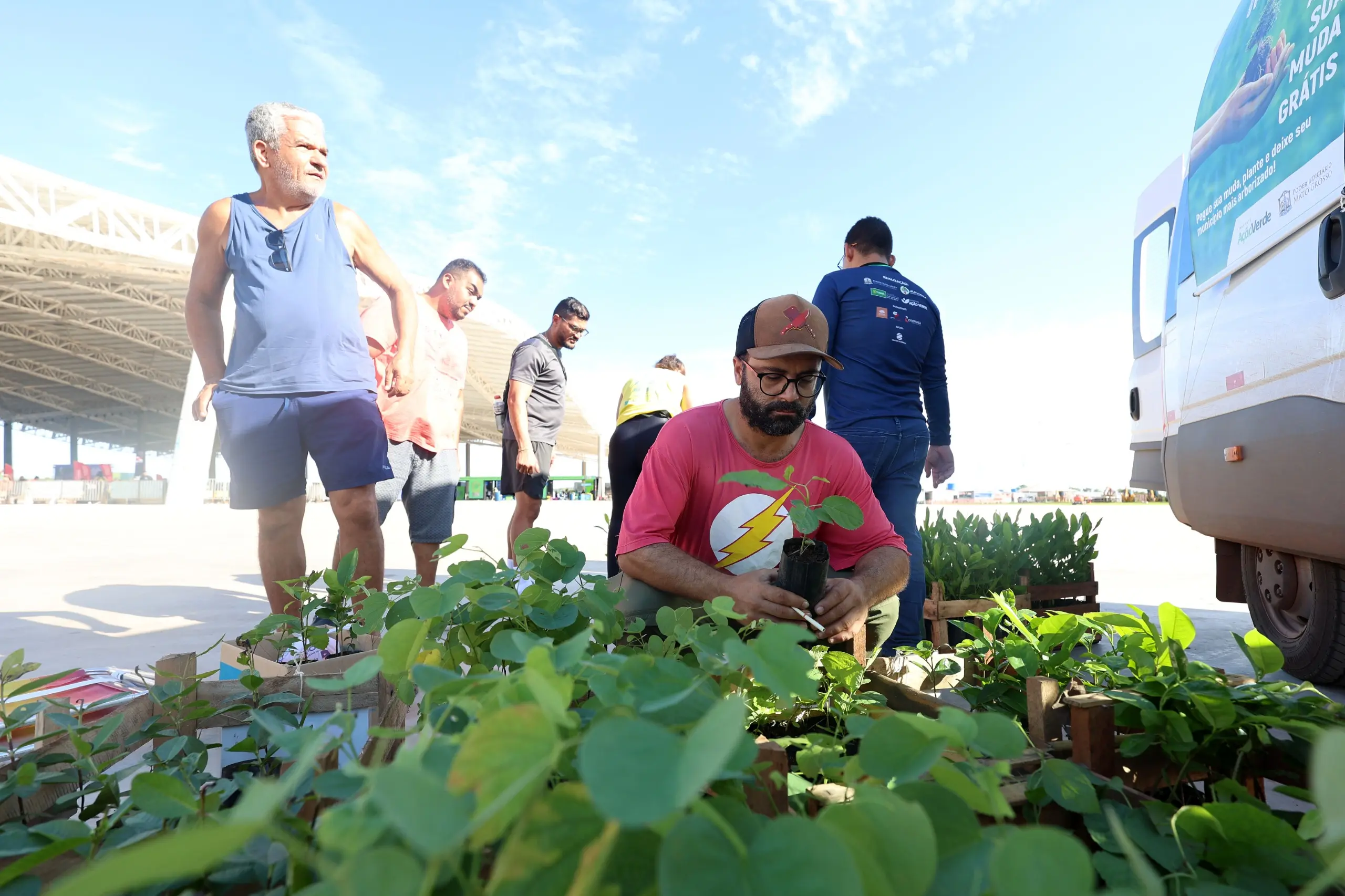 Homem agachado escolhe uma muda em caixas de madeira. Outros corredores observam e aguardam, em espaço aberto ao lado do veículo do projeto ambiental.