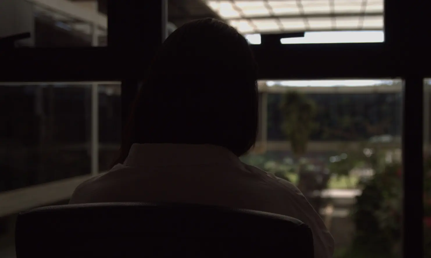 Foto horizontal que mostra a silhueta de uma mulher, a Luiza, sentada de costas. A luz da foto está escura para que ela não seja reconhecida.