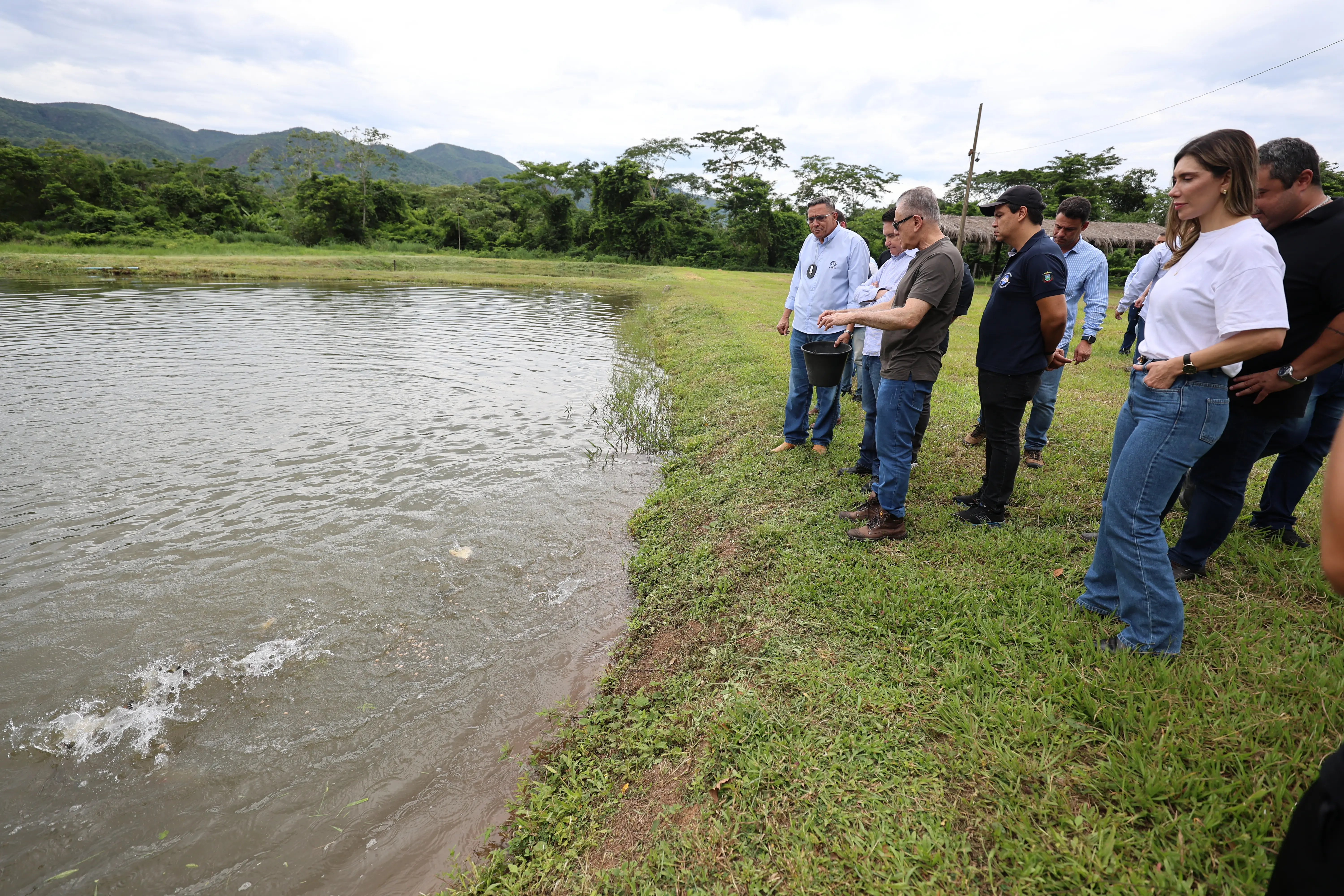 Um grupo de cerca de oito pessoas, incluindo homens e mulheres, vestindo roupas casuais, em pé na grama à beira de um lago. Um homem joga alimento na água. 