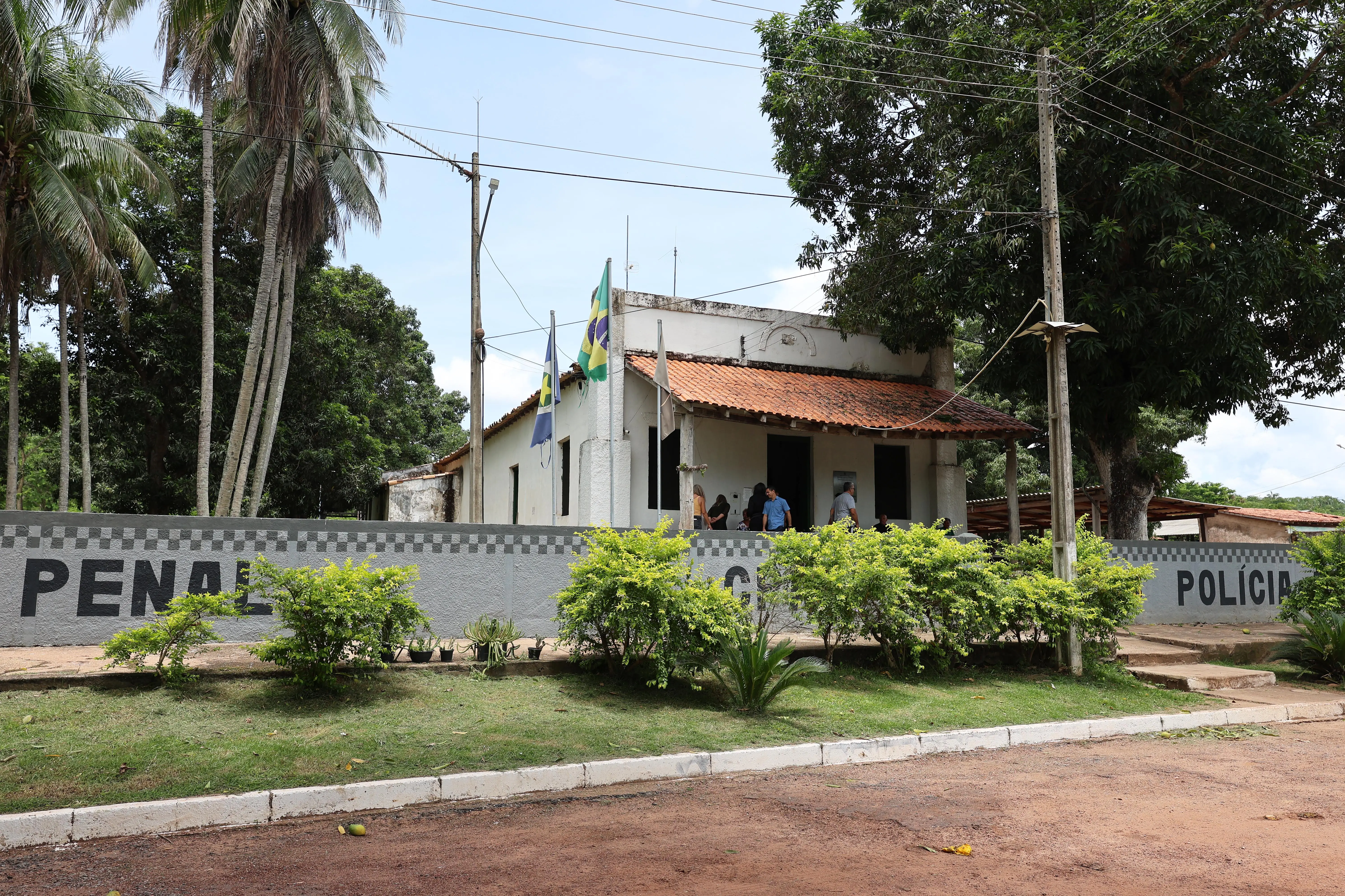 Vista frontal da fachada de um prédio antigo, branco com telhado de terracota, cercado por coqueiros e árvores. Há uma bandeira do Brasil na entrada e um muro baixo cinza.