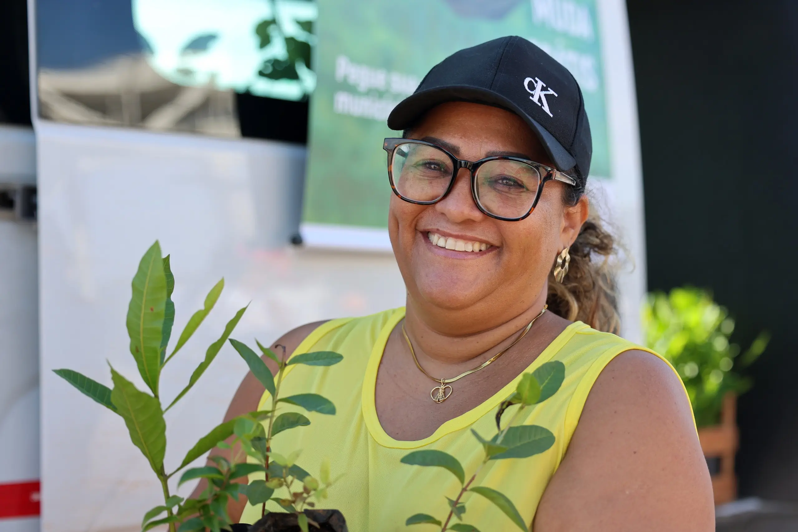 Mulher sorridente, usando boné e medalha, segura uma muda recém-recebida. A cena ocorre em área externa, com atletas e a van do projeto ao fundo.